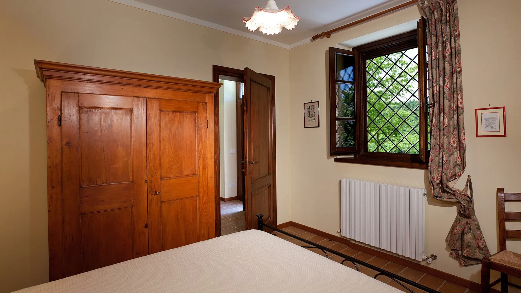 A bedroom with a large wooden wardrobe and a window overlooking greenery.
