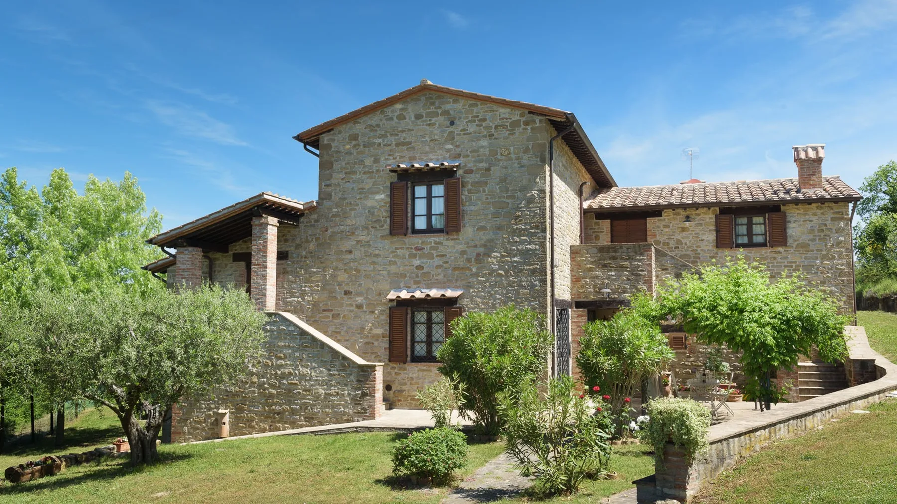 A stone house with a tiled roof stands among trees and garden shrubs.