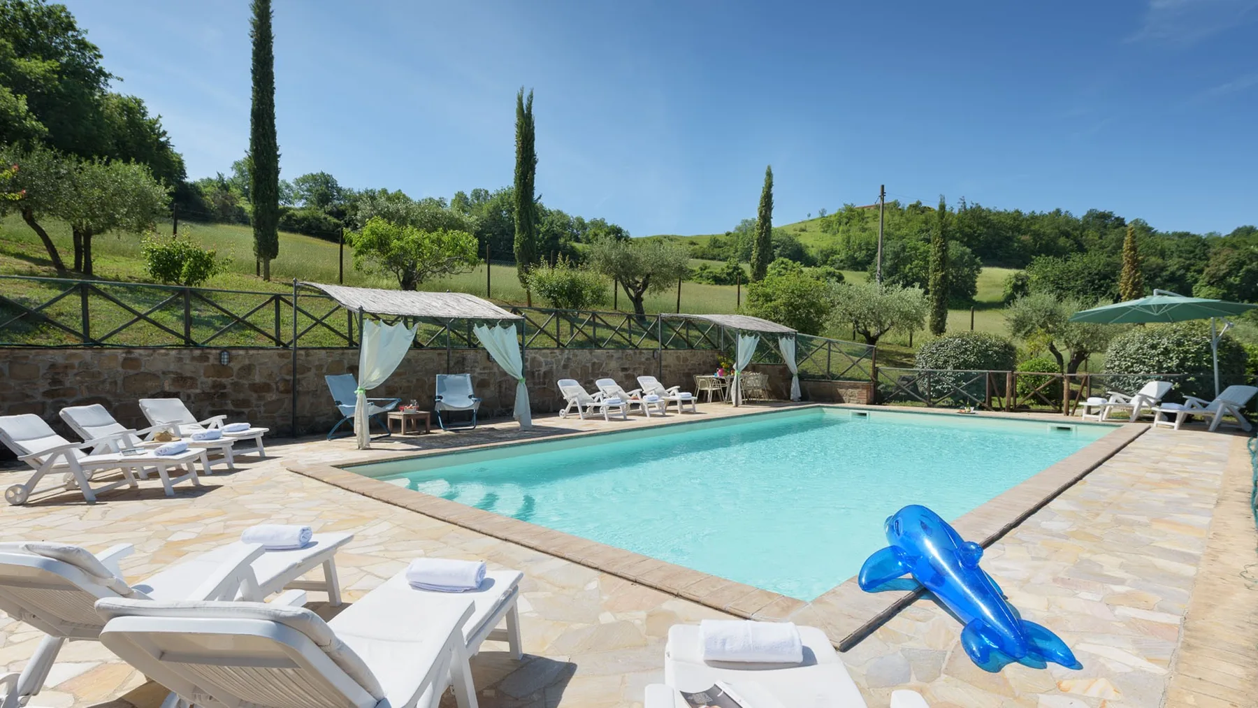 An outdoor swimming pool sits beside a paved deck with lounge chairs and a blue inflatable dolphin.