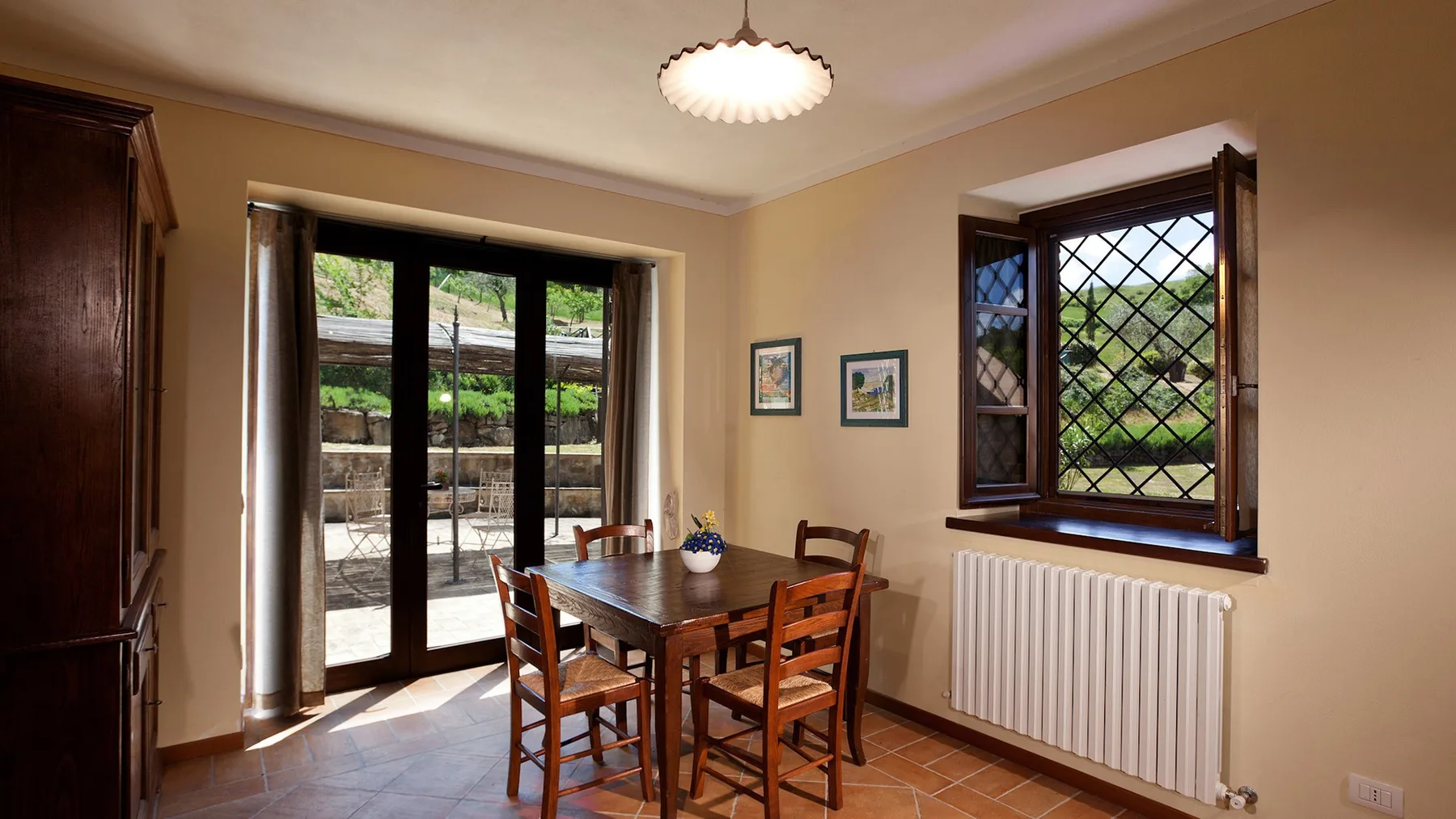 A dining area with a wooden table and chairs beside windows overlooking greenery.