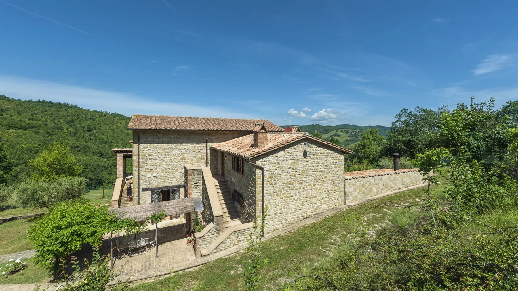 A stone building with tiled roofs sits among green hills under a blue sky.