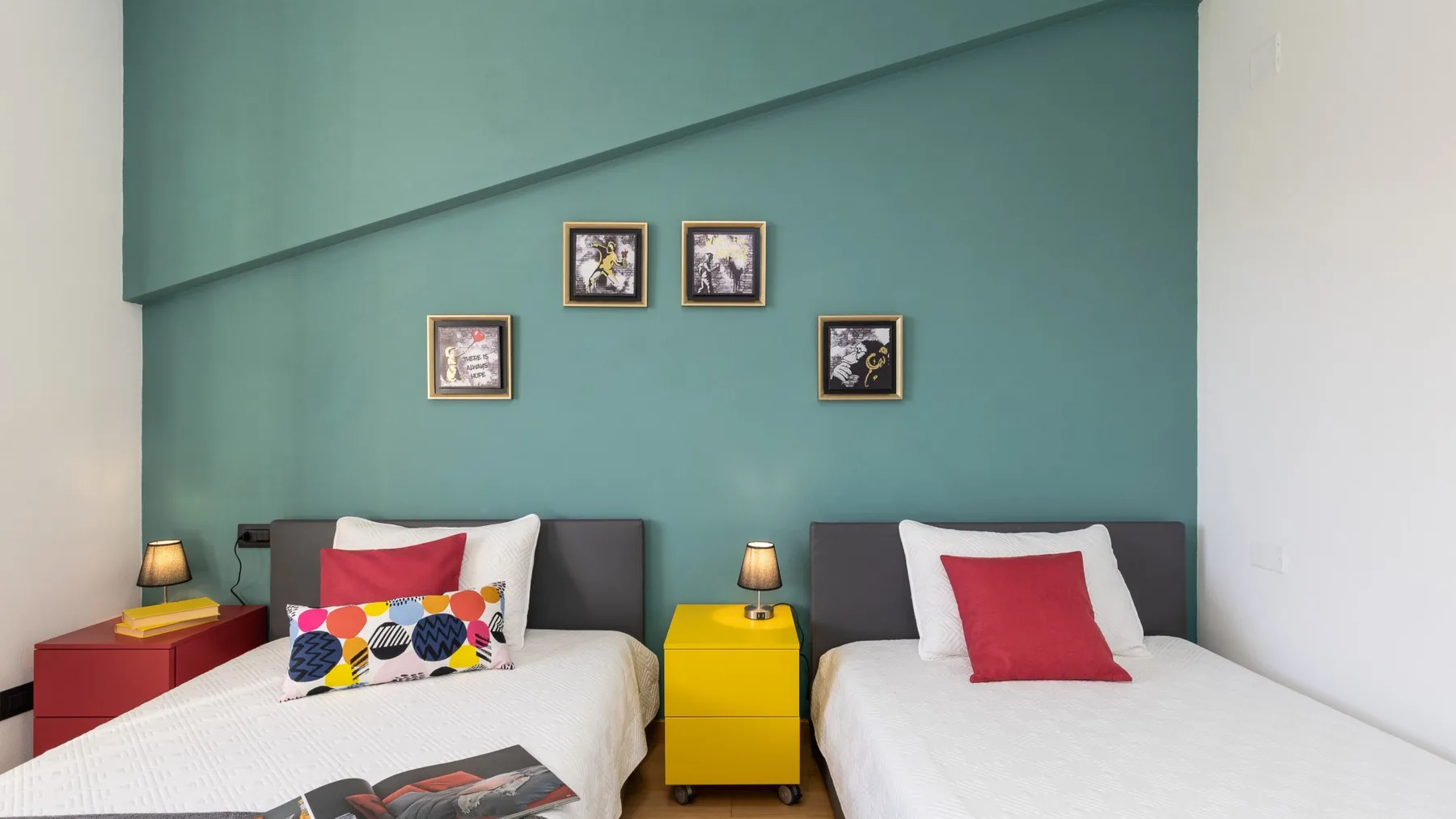 A bedroom with two beds, colorful pillows, and framed pictures on a teal wall.