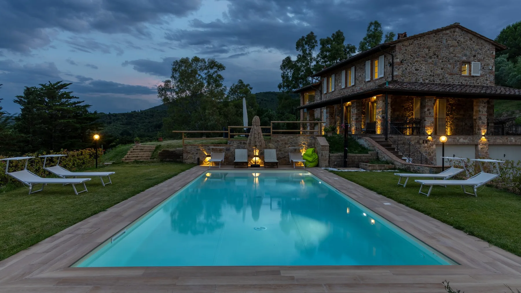 An outdoor swimming pool beside a stone house, with lawn seating and a cloudy evening sky.