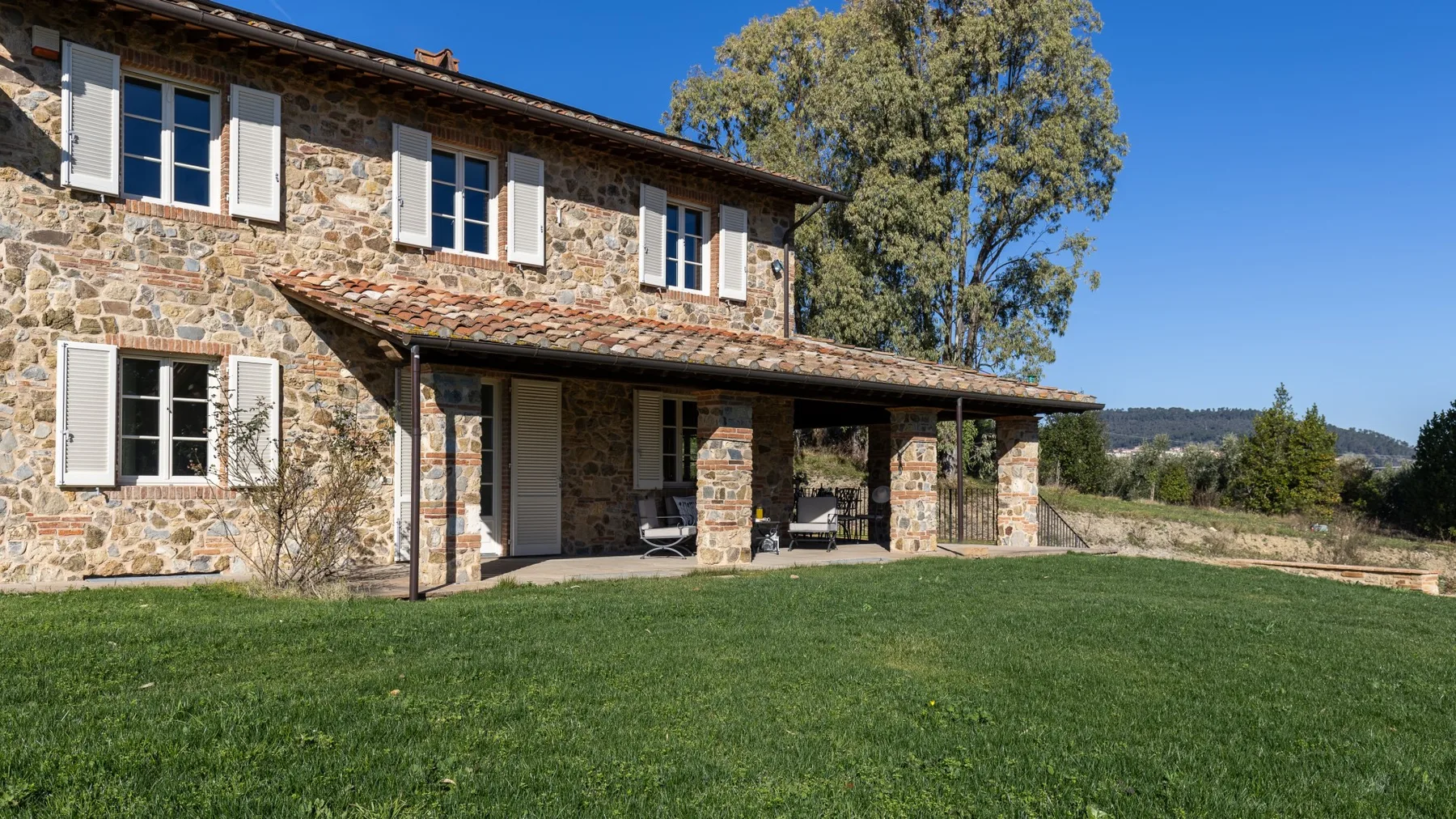 A stone house with shuttered windows and a covered porch faces a broad green lawn.