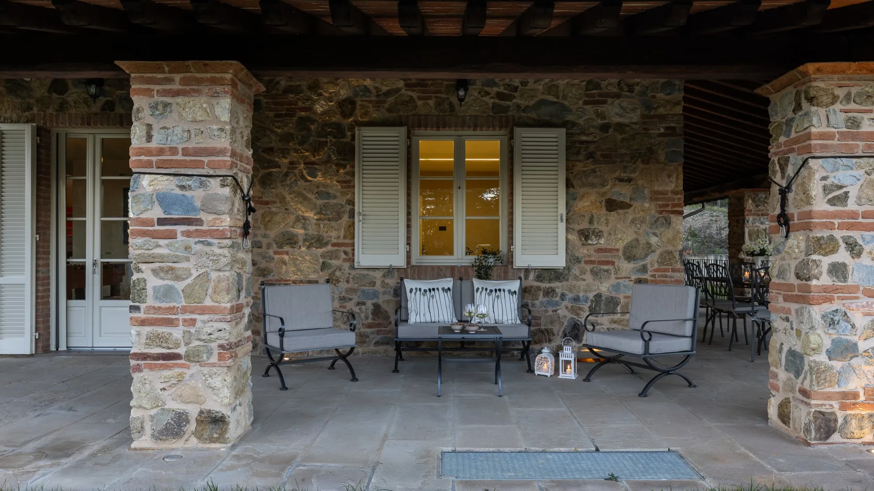 A covered stone patio has cushioned seating around a low table.