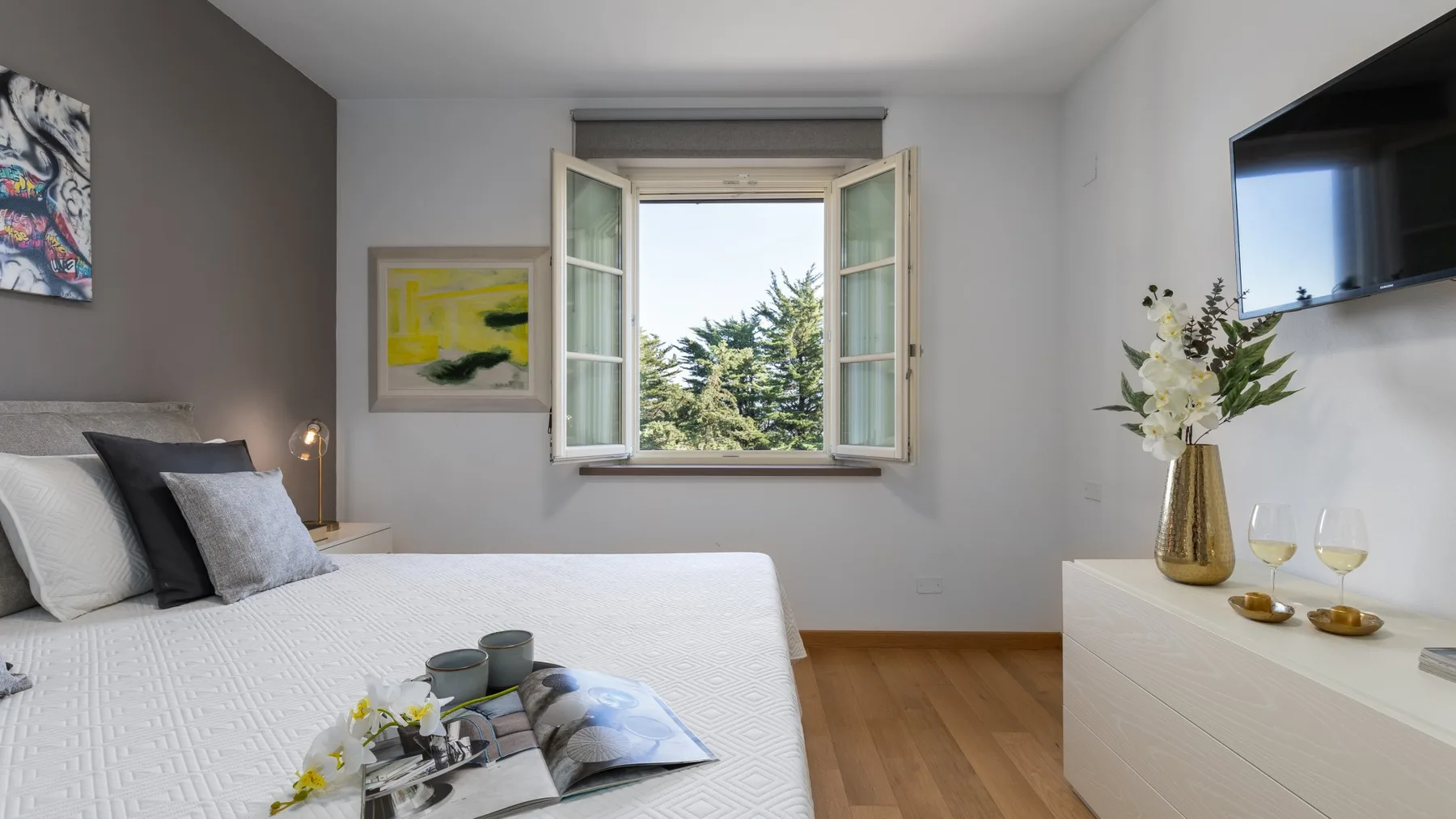 A bright bedroom with a neatly made bed and an open window overlooking trees.