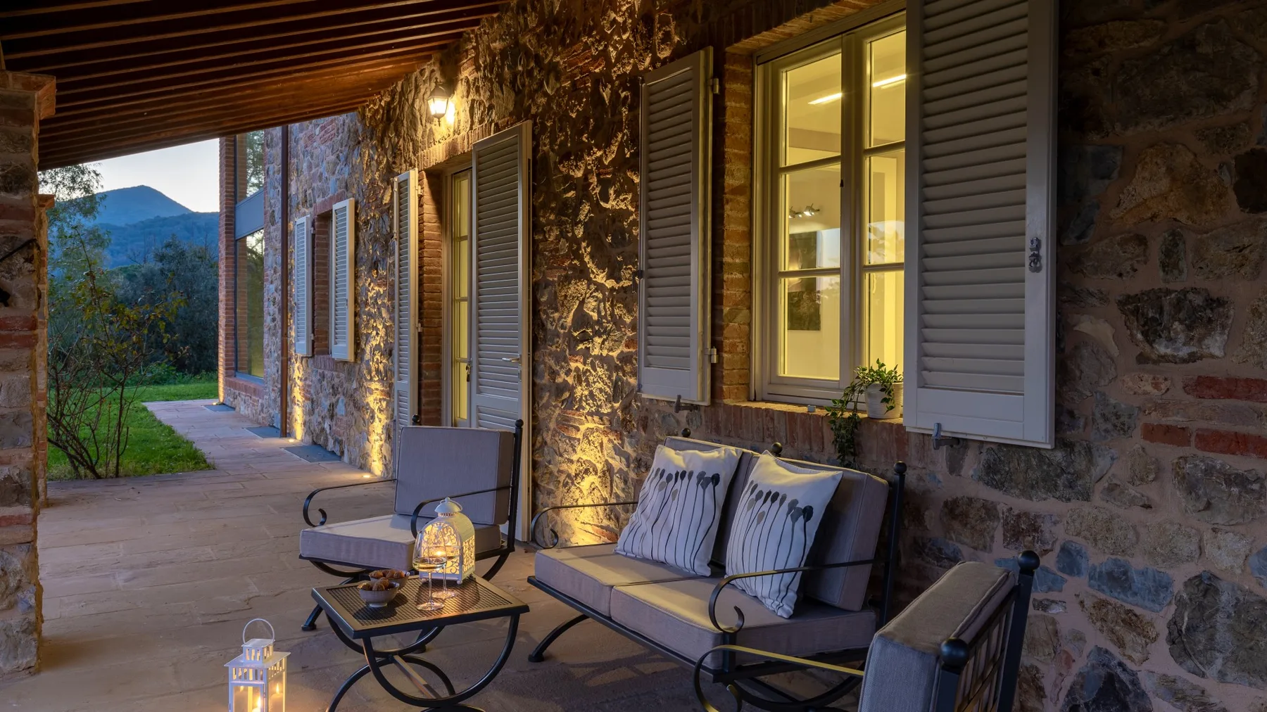 A covered stone veranda has outdoor seating beside warmly lit windows.