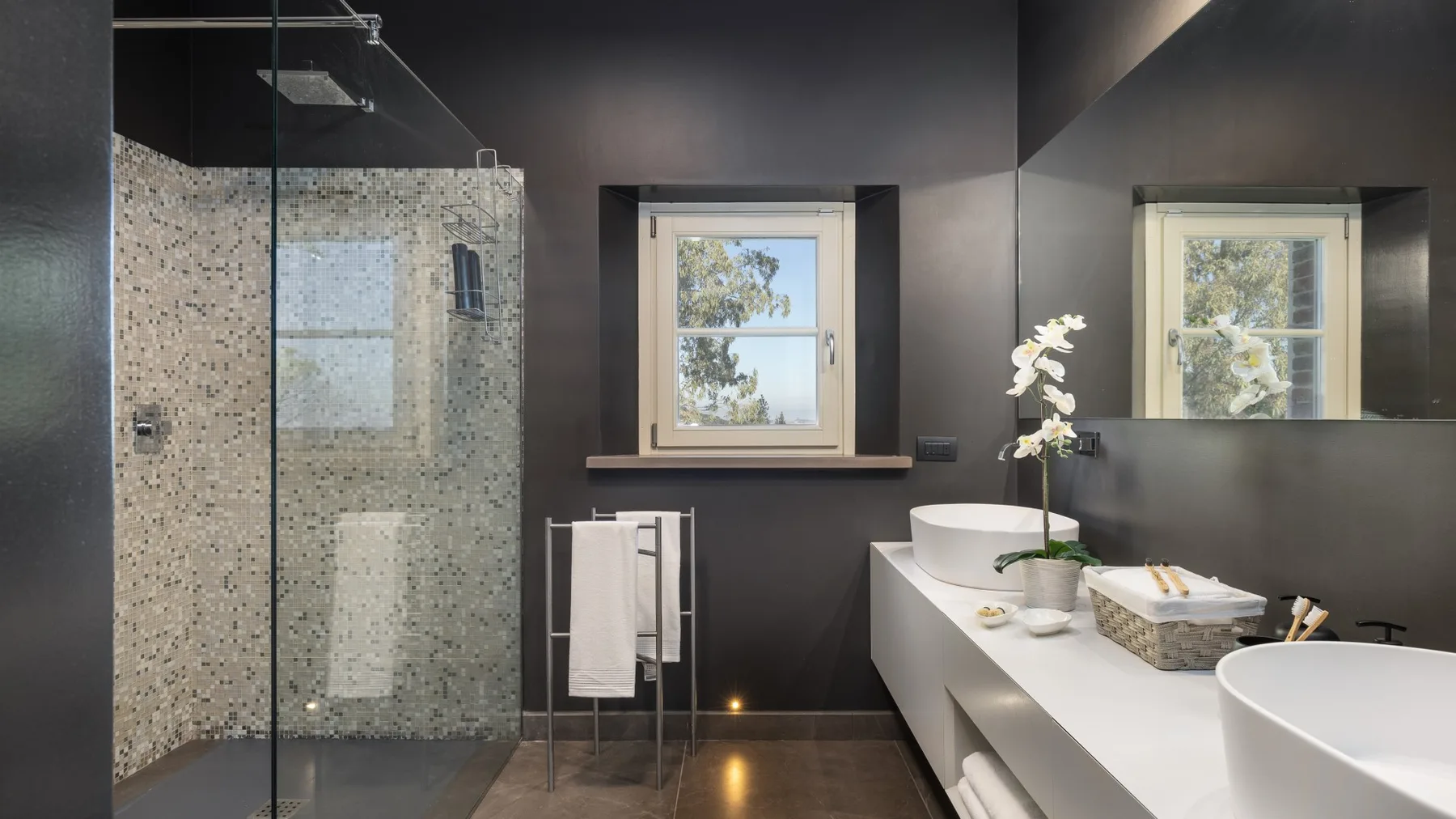 A modern bathroom with a glass shower, a long vanity, and two windows.