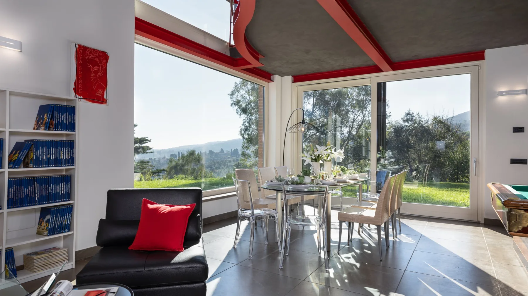 A bright dining area with a glass table and large windows overlooking greenery.