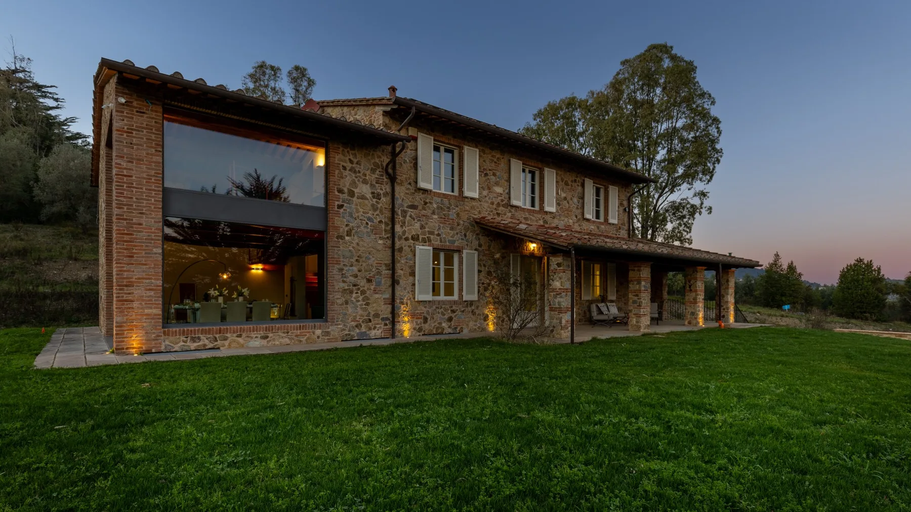 A stone-and-brick house with a large illuminated window faces a grassy lawn.