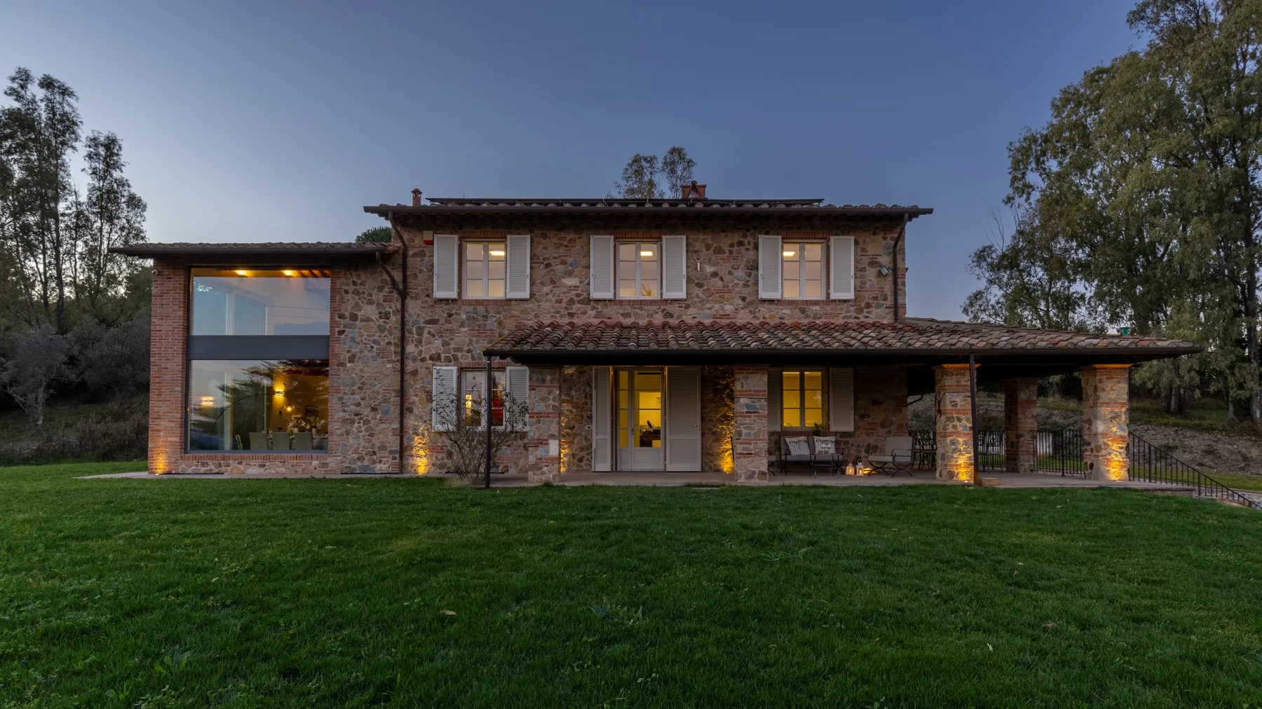 A stone-faced two-story house stands beyond a broad green lawn at dusk.