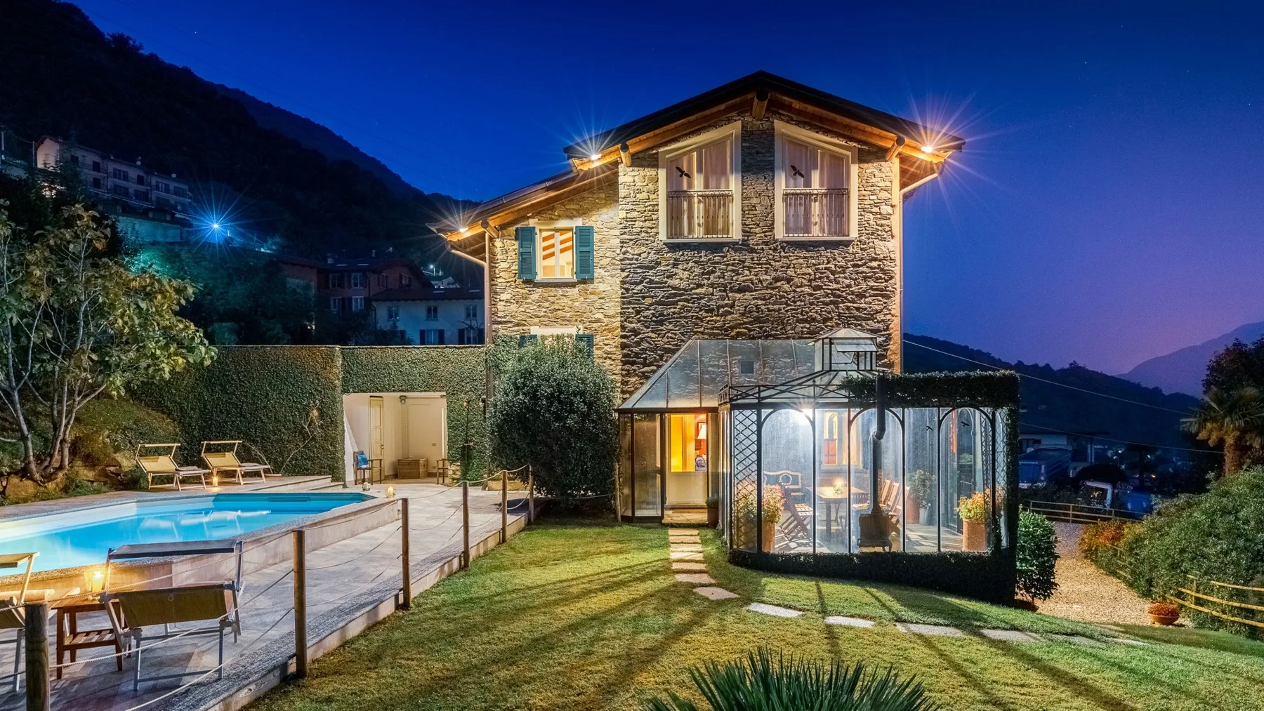 Una casa de piedra con un patio acristalado está junto a una piscina iluminada al anochecer.