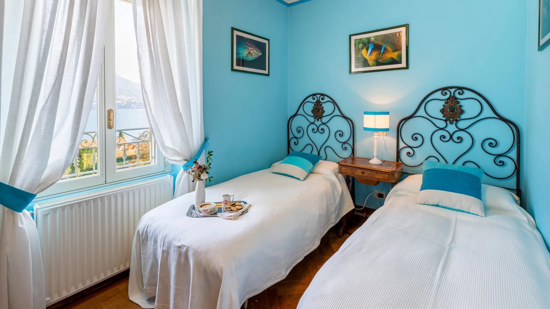 Un dormitorio luminoso con dos camas, paredes turquesas y una ventana con cortinas transparentes.