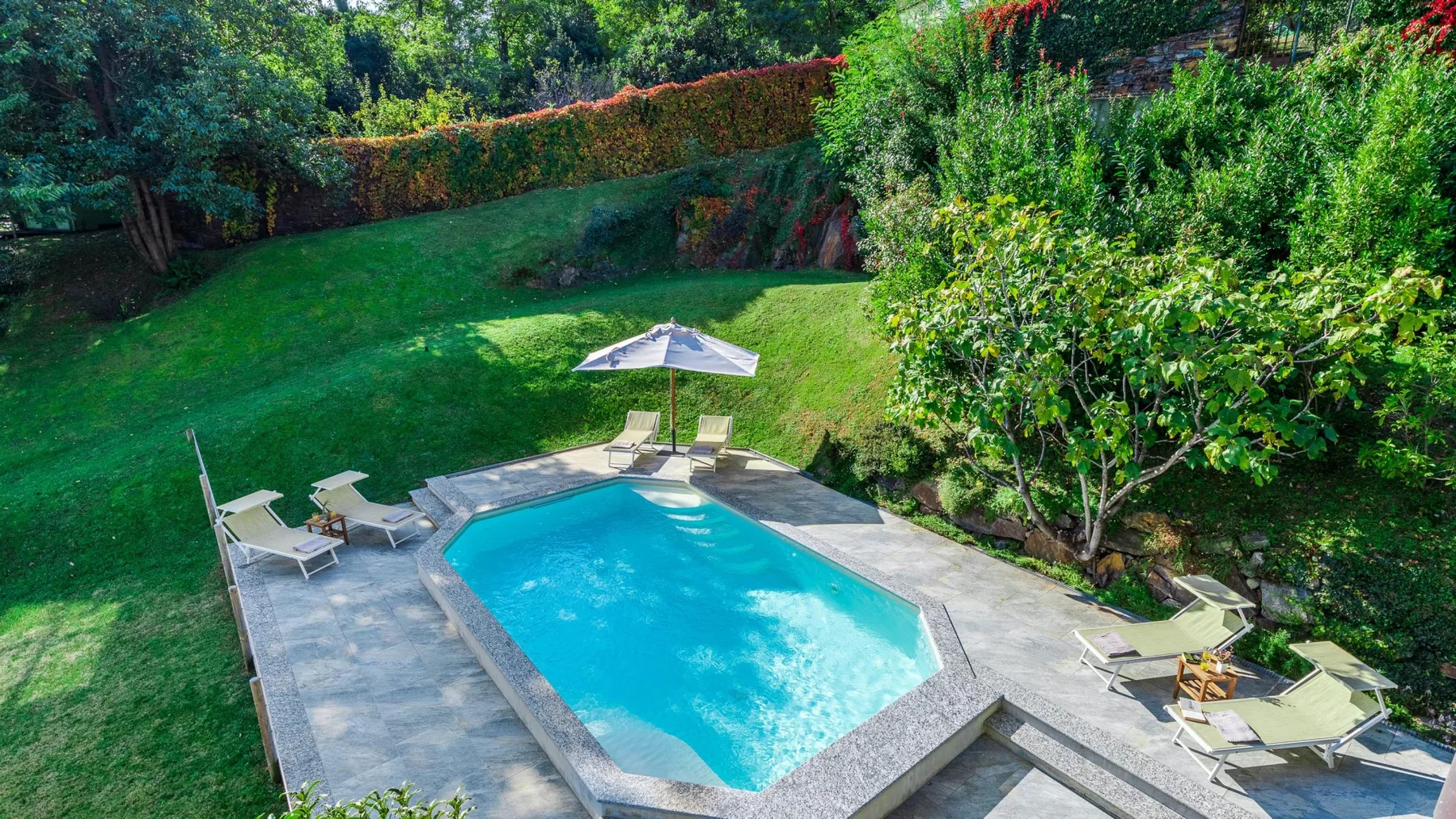 Una piscina al aire libre está junto a un césped y un jardín frondoso.