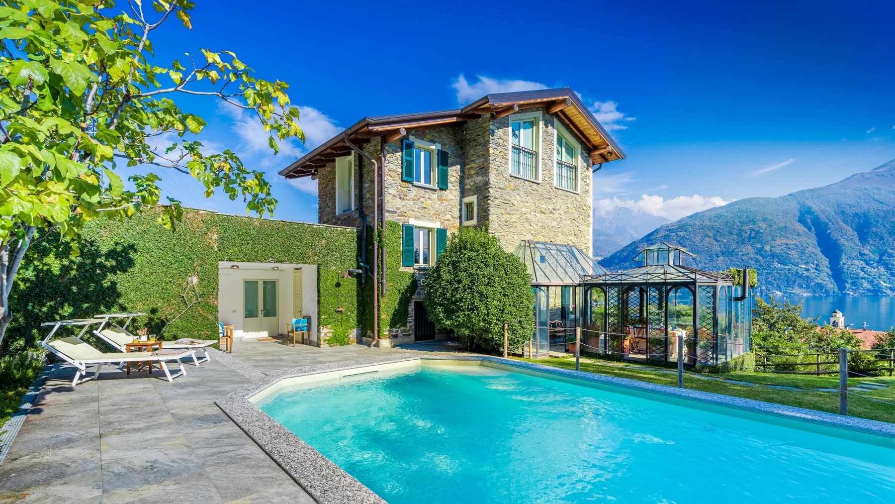 Una casa de piedra está junto a una piscina turquesa, con montañas al fondo.