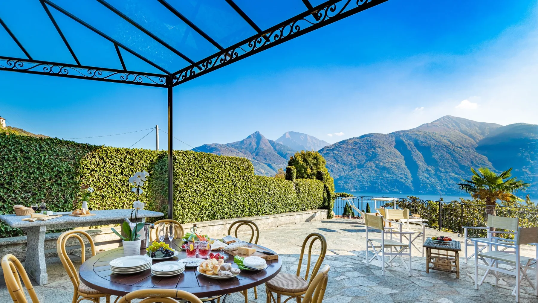 Terraza junto al lago con asientos al aire libre y vistas a las montañas bajo una estructura metálica.