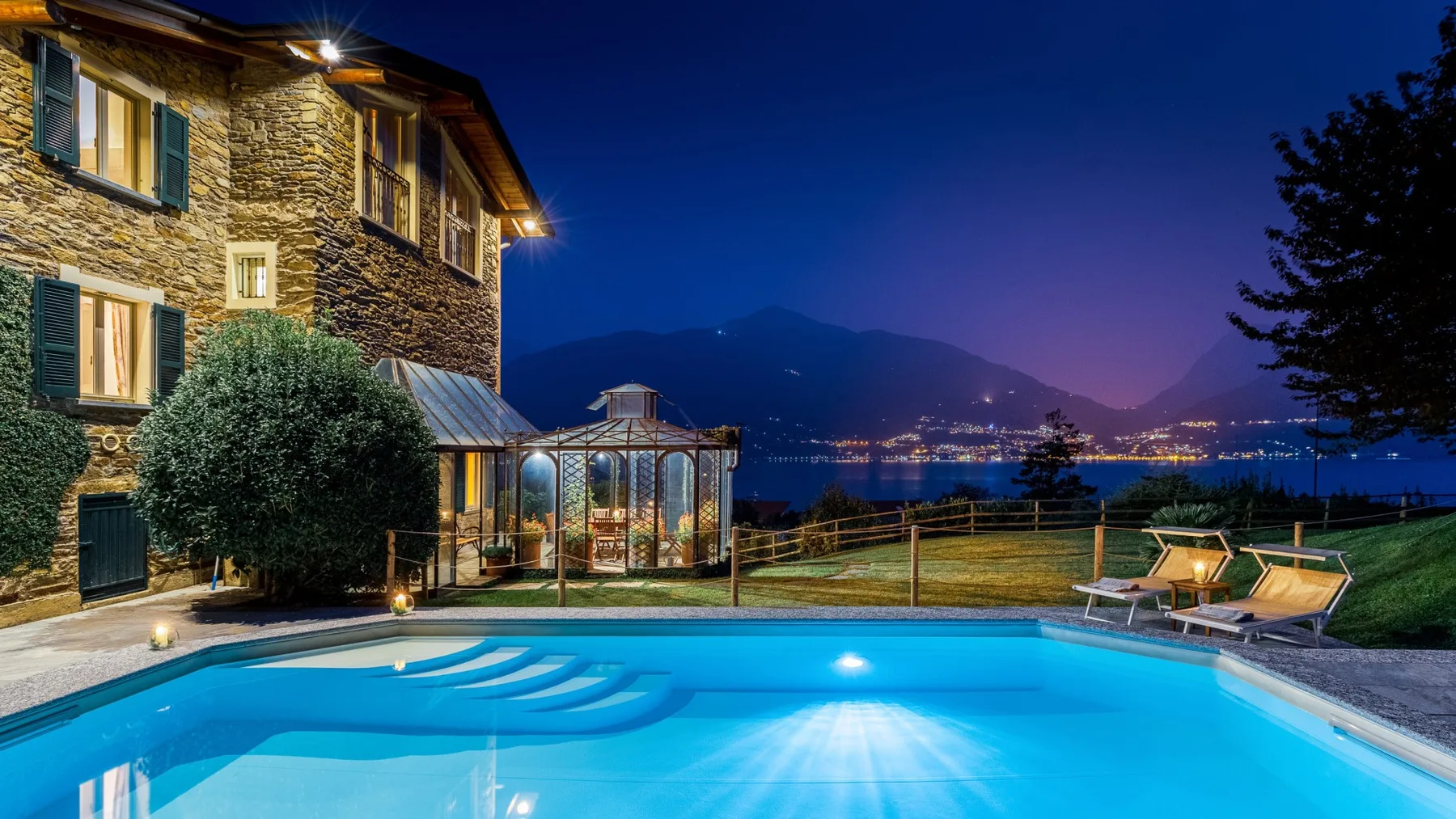 Una piscina al aire libre brilla junto a un edificio de piedra al anochecer, con montañas al fondo.