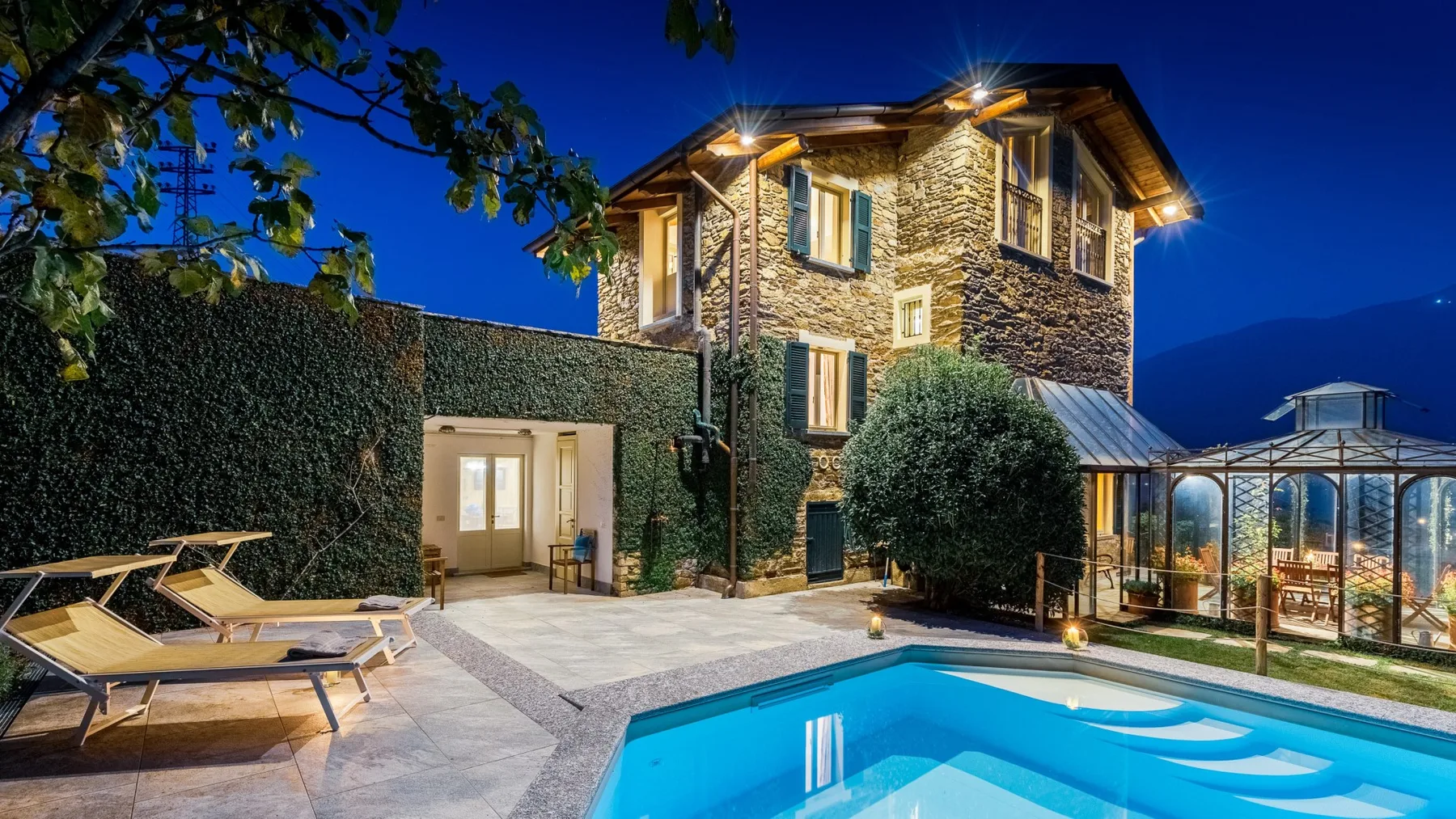 Una casa de piedra junto a una piscina iluminada y un patio acristalado por la noche.