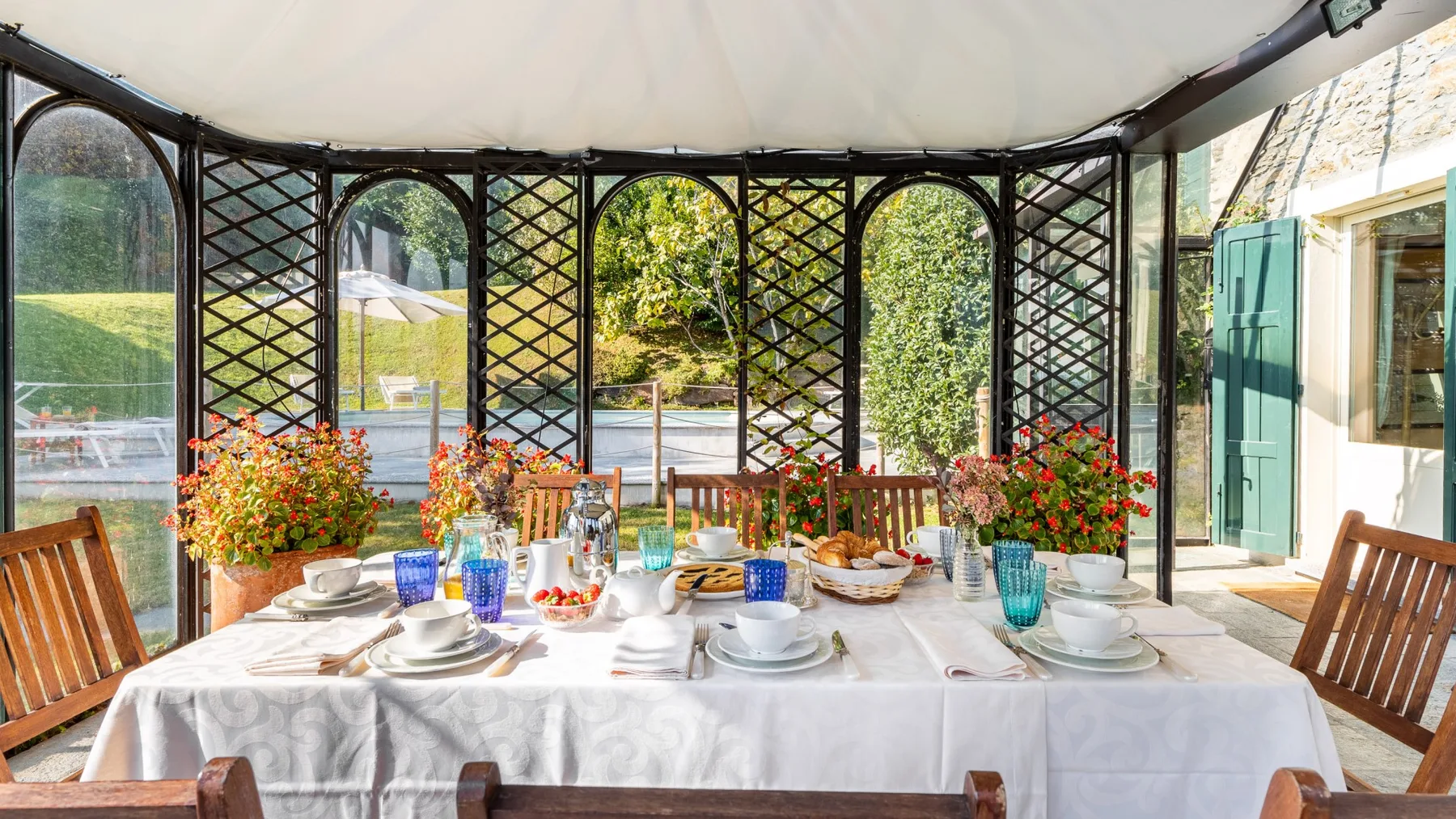 Un comedor luminoso con una mesa larga preparada junto a paneles de vidrio arqueados y plantas con flores.
