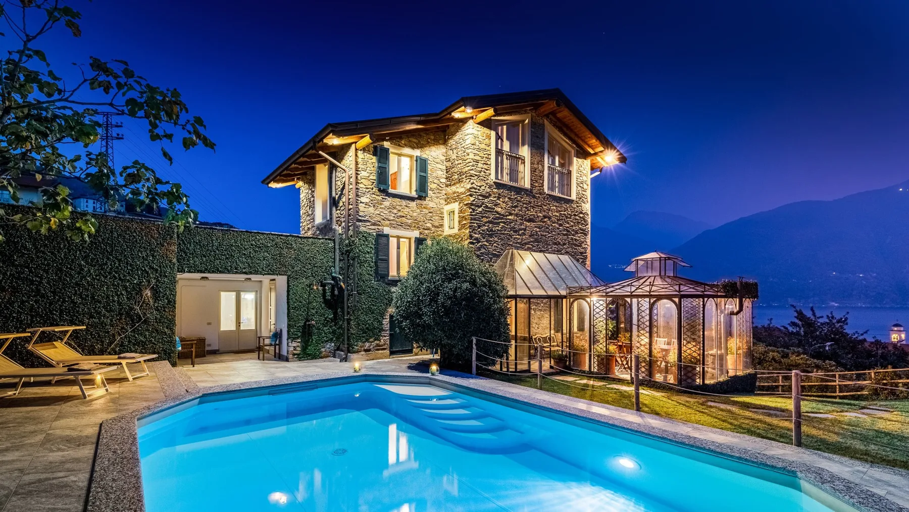 Una casa de piedra iluminada da a una piscina y un invernadero acristalado al anochecer.