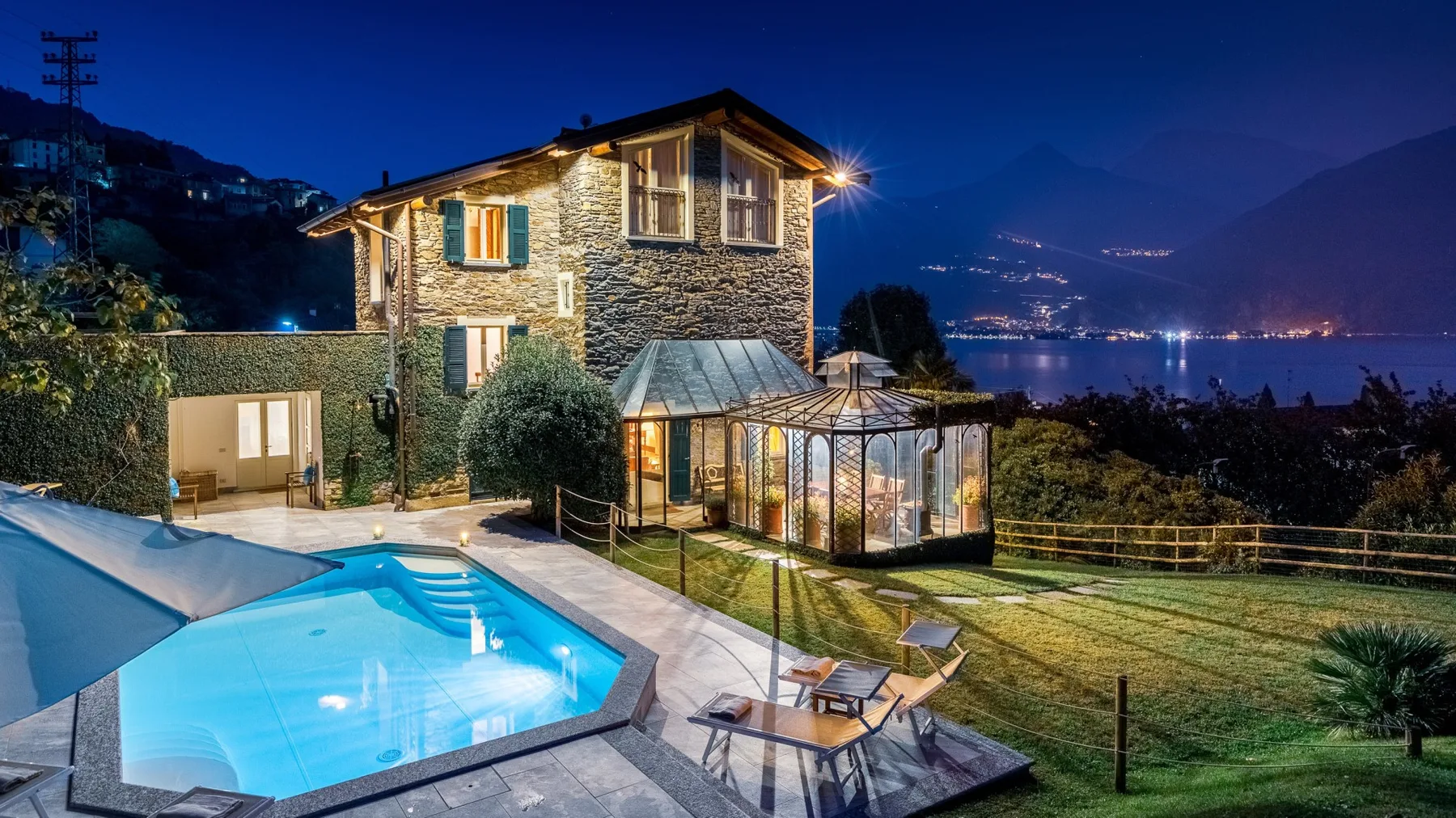Una casa de piedra y una piscina iluminada miran hacia un paisaje junto al lago por la noche.