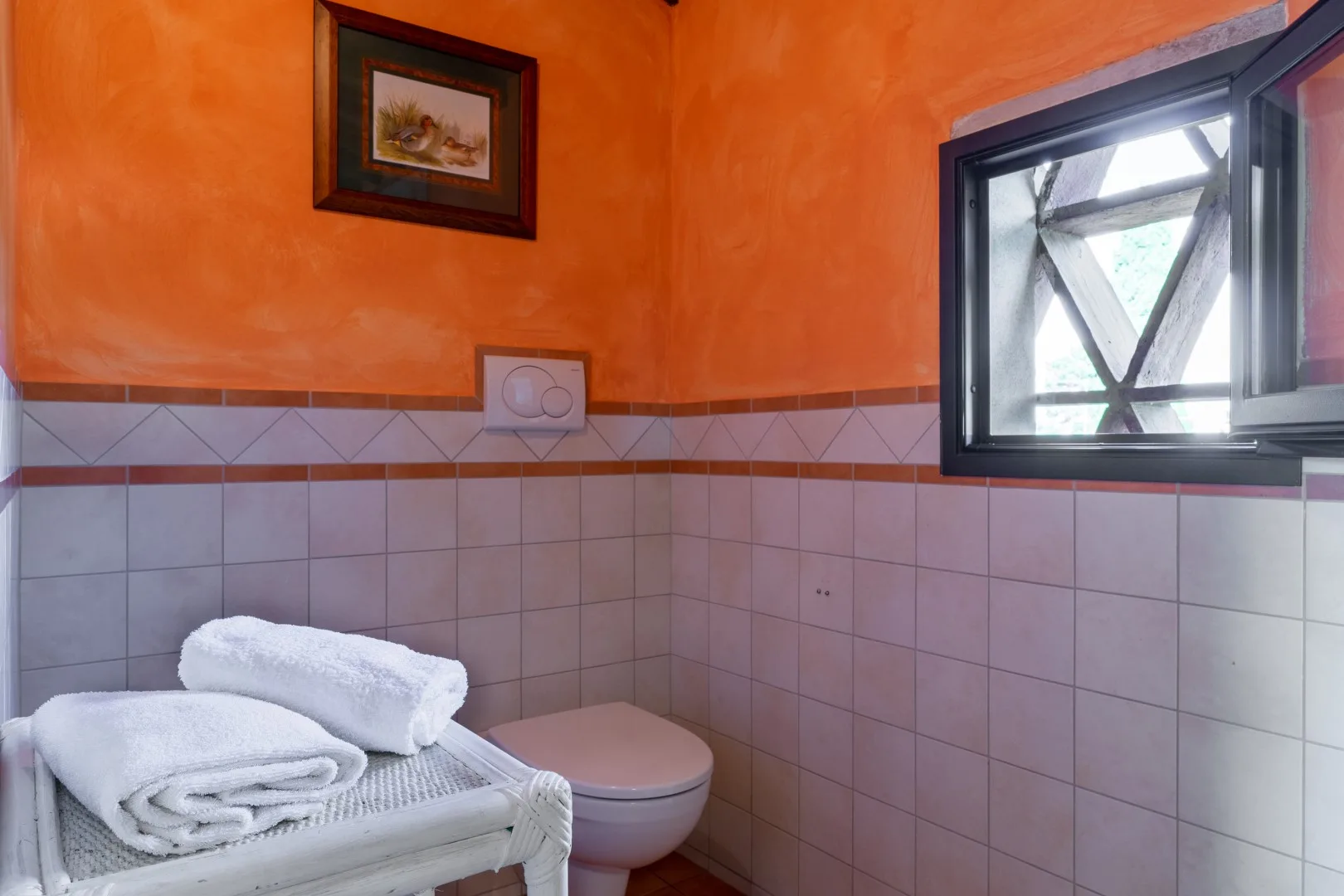 Un baño alicatado tiene un inodoro blanco, toallas dobladas, paredes superiores naranjas y una ventana abierta.