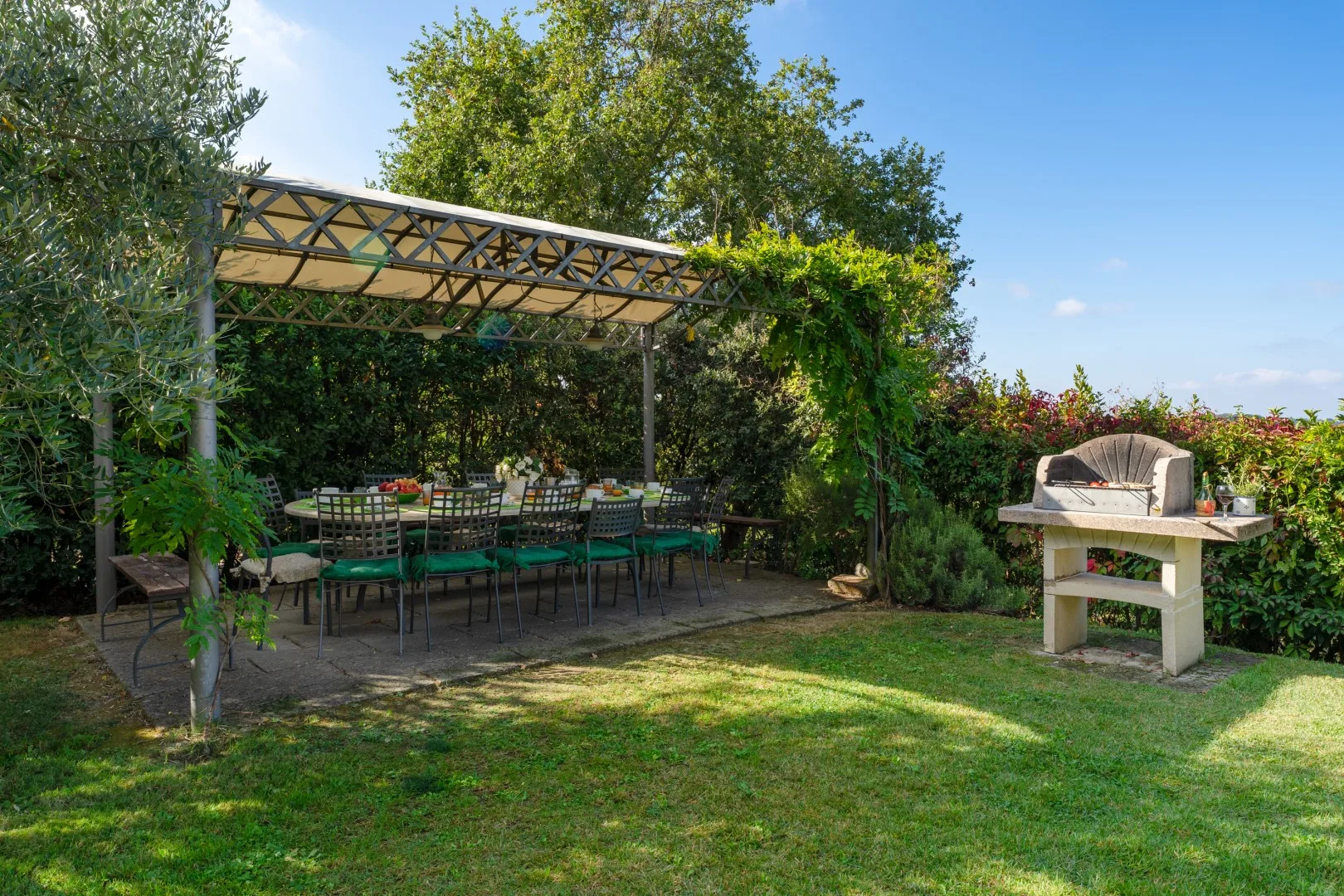 Un patio ajardinado tiene una mesa de comedor bajo una pérgola y una barbacoa de obra cerca.