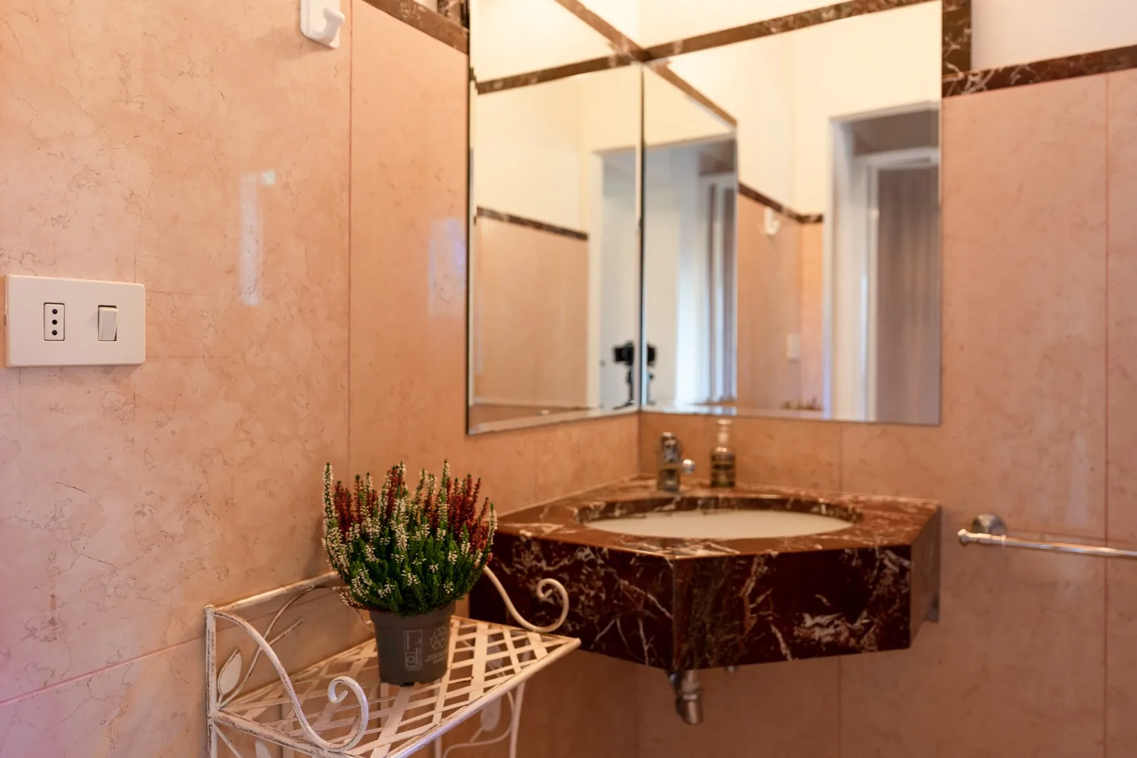 Un lavabo de baño revestido de azulejos tiene una encimera de mármol oscuro, un espejo y una planta en un soporte pequeño.