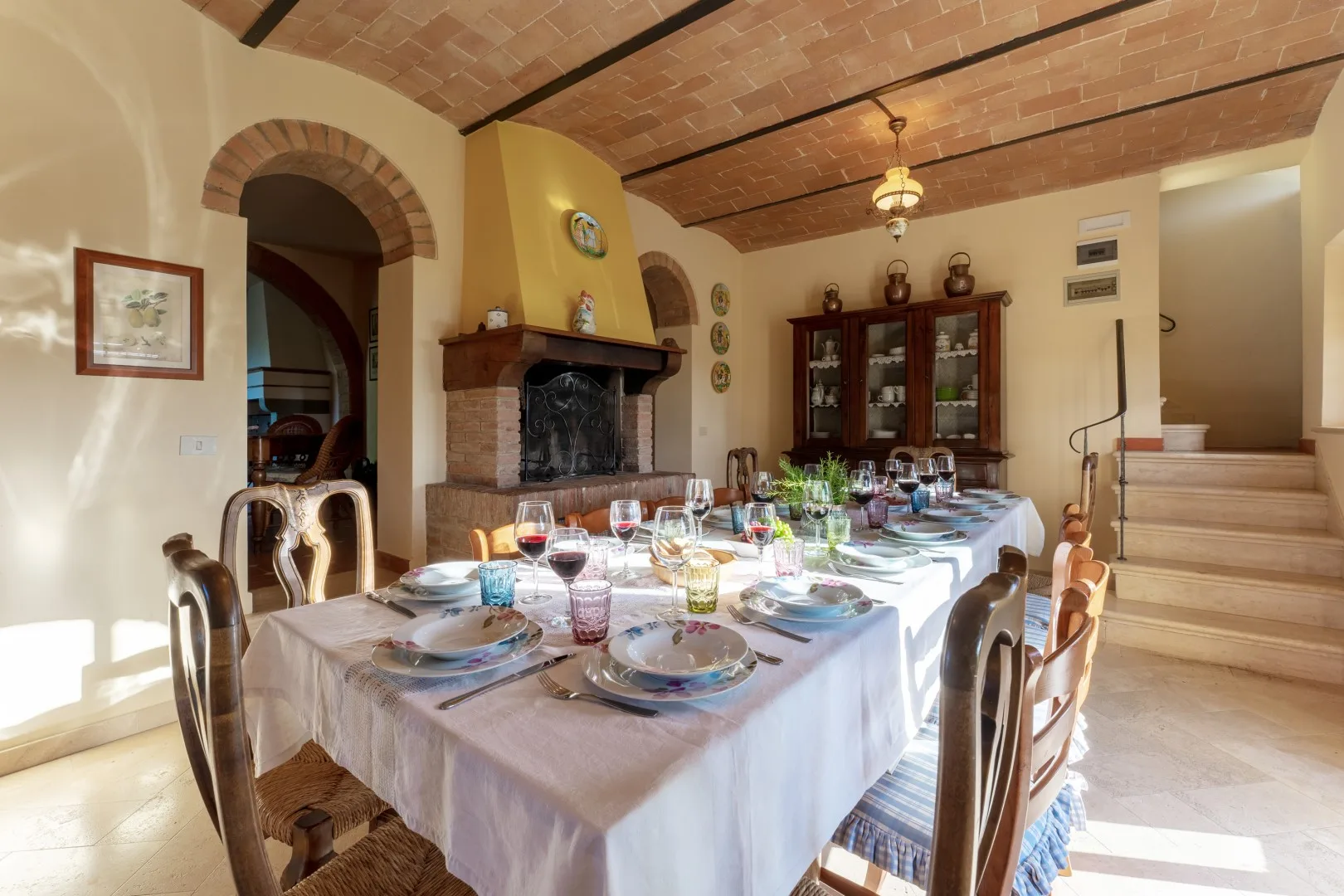Un comedor con una larga mesa preparada con platos, vasos y fuentes bajo un techo de ladrillo.