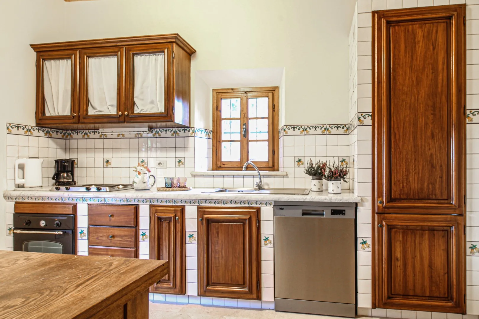 Una cocina alicatada cuenta con armarios de madera, una encimera, un fregadero bajo una ventana y un lavavajillas de acero inoxidable.