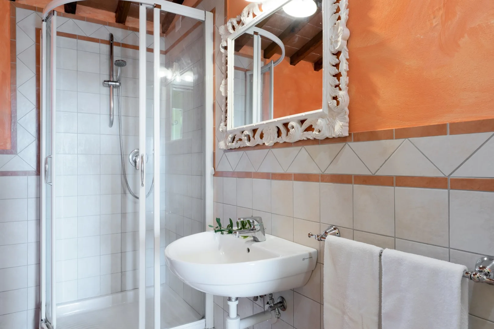 Baño con ducha acristalada, lavabo blanco y toallas bajo un espejo ornamentado.