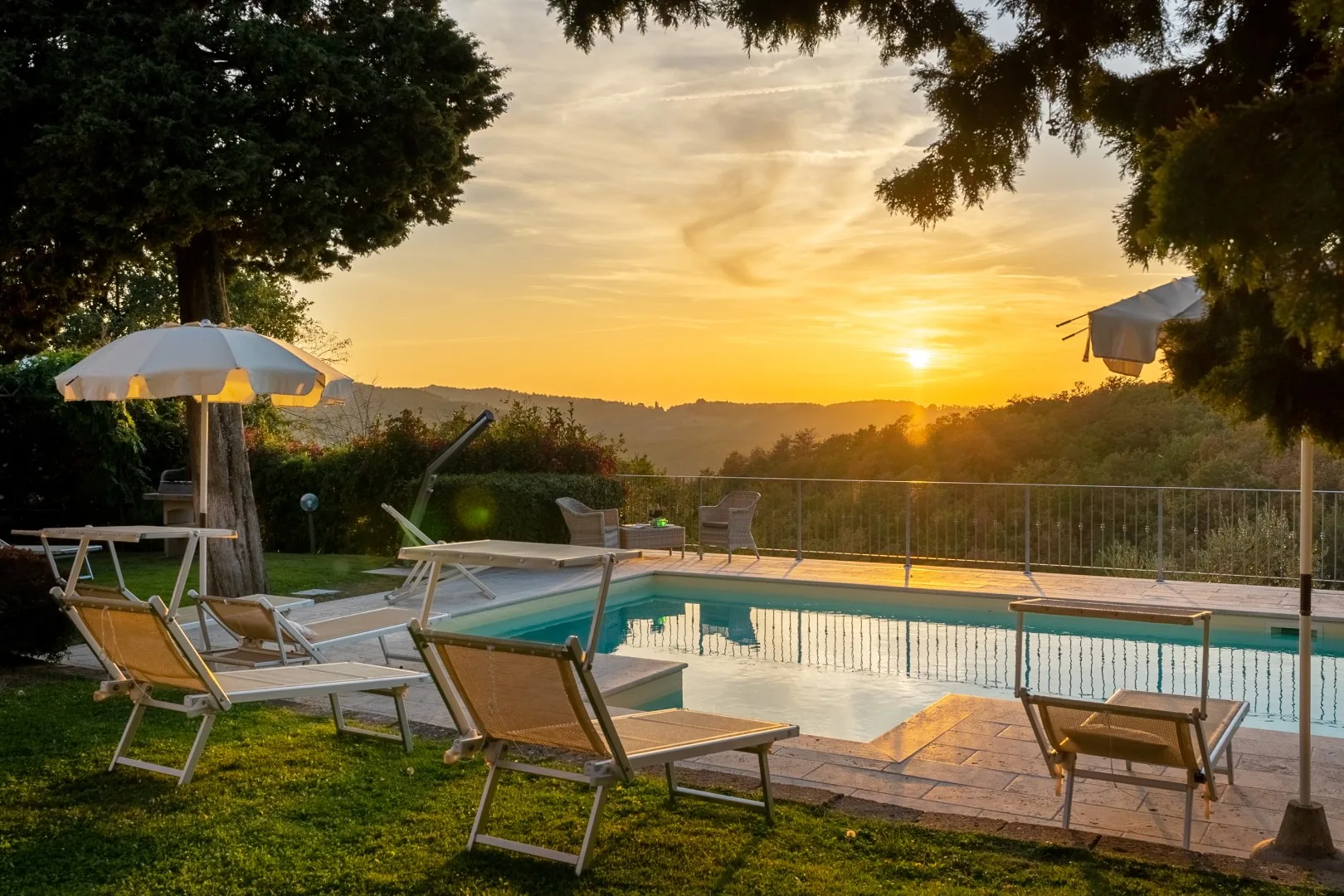 Una piscina y tumbonas miran hacia colinas lejanas al atardecer, enmarcadas por árboles y una sombrilla abierta.