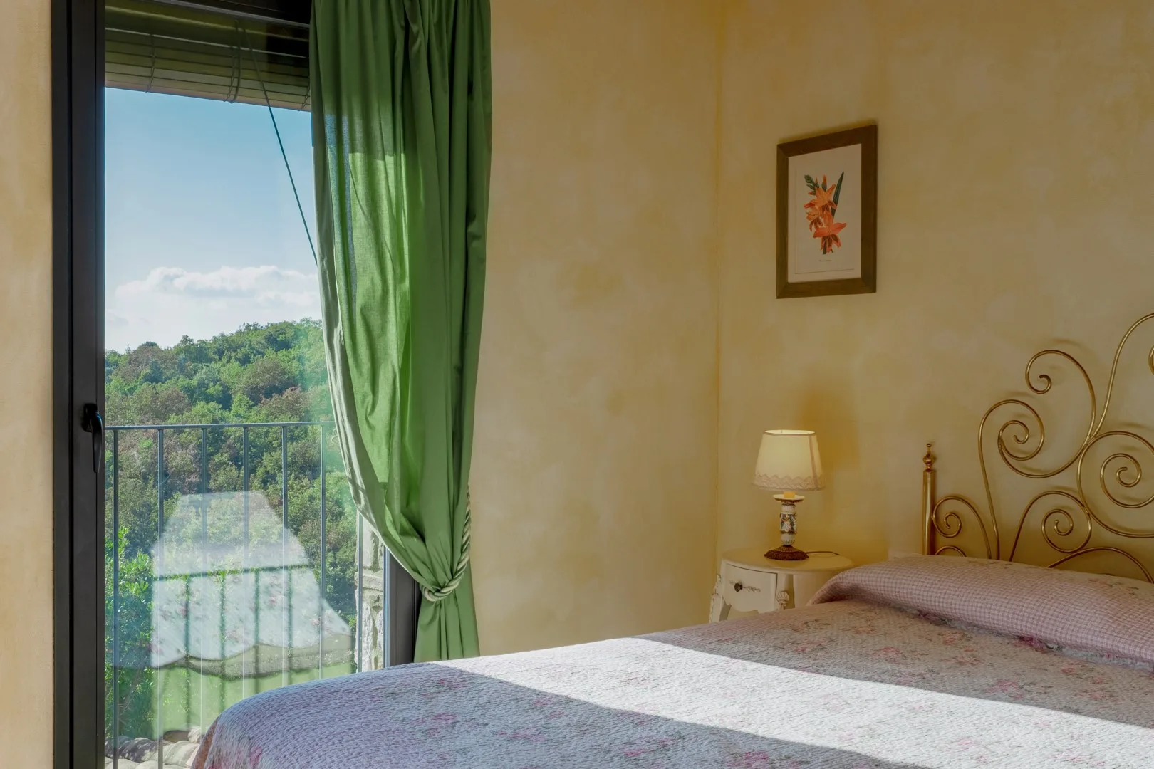 Dormitorio con una colcha estampada, un cabecero metálico ornamentado y cortinas verdes junto a una puerta al balcón.