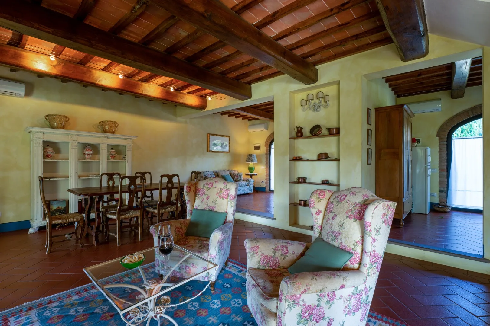Una cálida sala de estar con vigas de madera vistas, sillones florales, una mesa de centro de vidrio y un comedor.