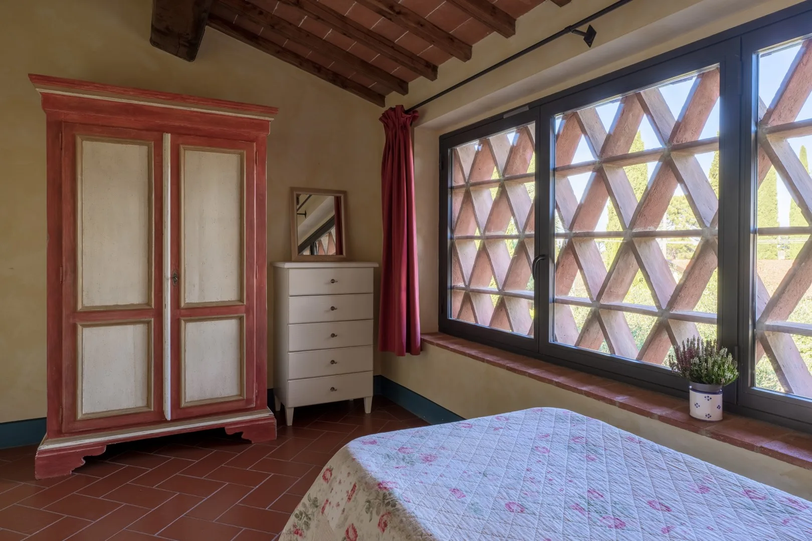 Un dormitorio con un armario de molduras rojas, una cómoda y una cama junto a grandes ventanas geométricas.