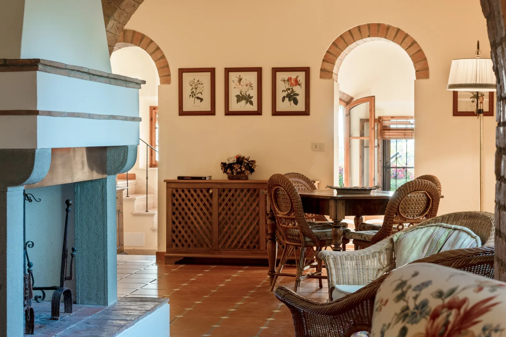 Una sala de estar en tonos cálidos con chimenea de azulejos, sillas de mimbre, mesa de madera y láminas botánicas enmarcadas.