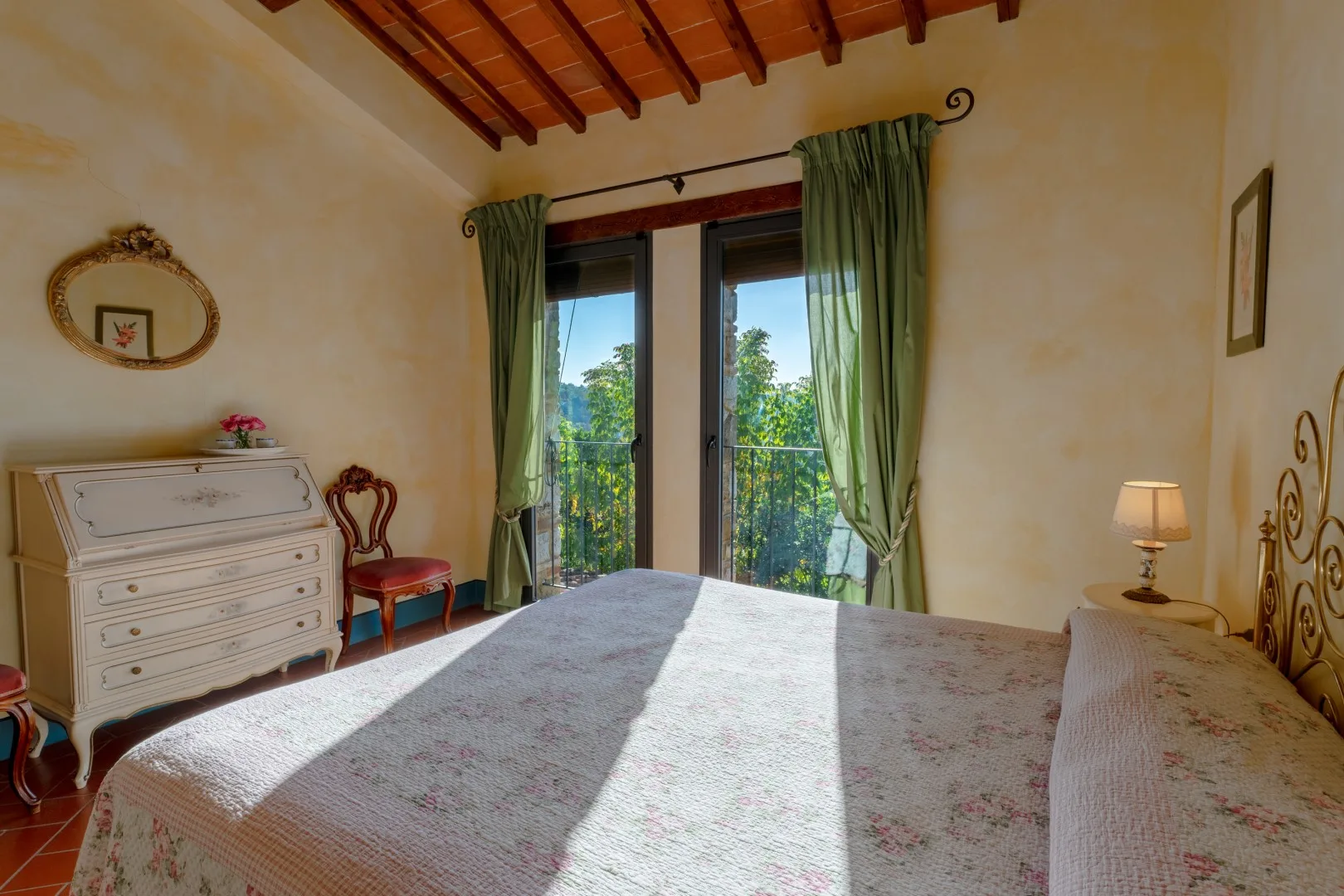 Un dormitorio con una cama estampada, una cómoda blanca, cortinas verdes y puertas de vidrio que dan a un balcón.