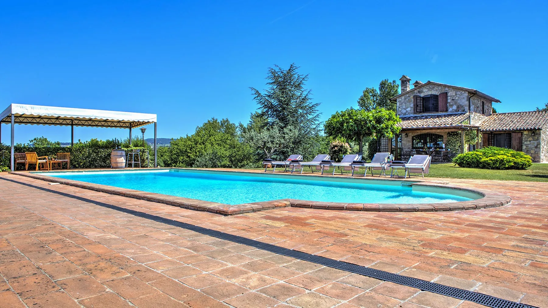 A swimming pool sits beside a broad terracotta-paved patio, with loungers, a canopy, and a stone house beyond.
