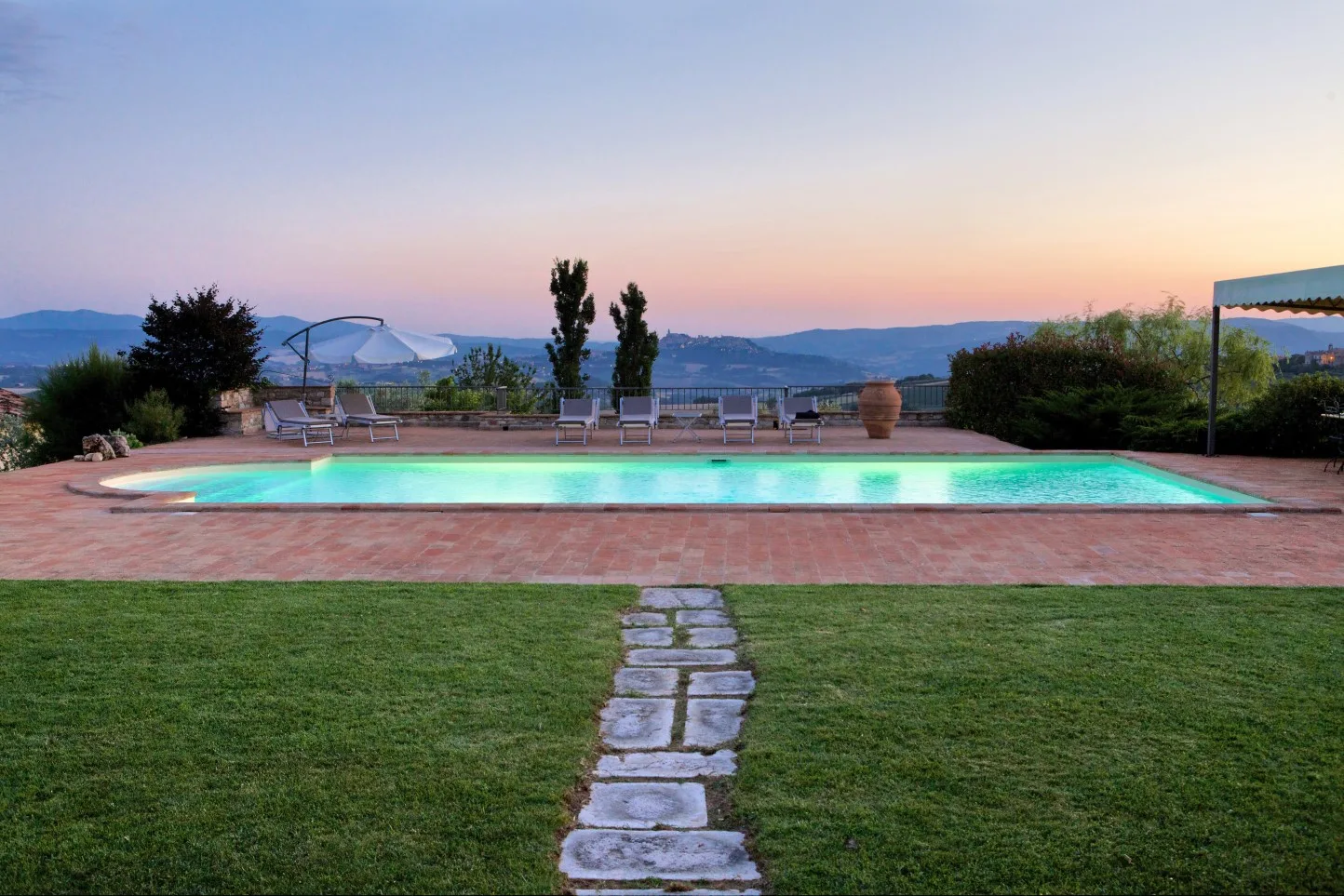 Outdoor pool scene with a stone path crossing the lawn, lounge chairs, and hills at dusk.