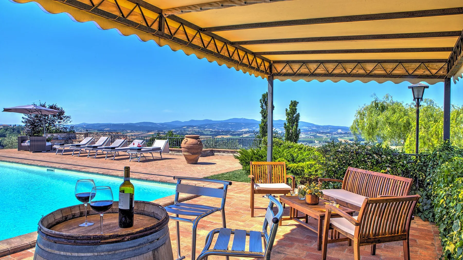 A covered terrace overlooks a swimming pool, with wooden seating, a wine barrel table, and distant hills.