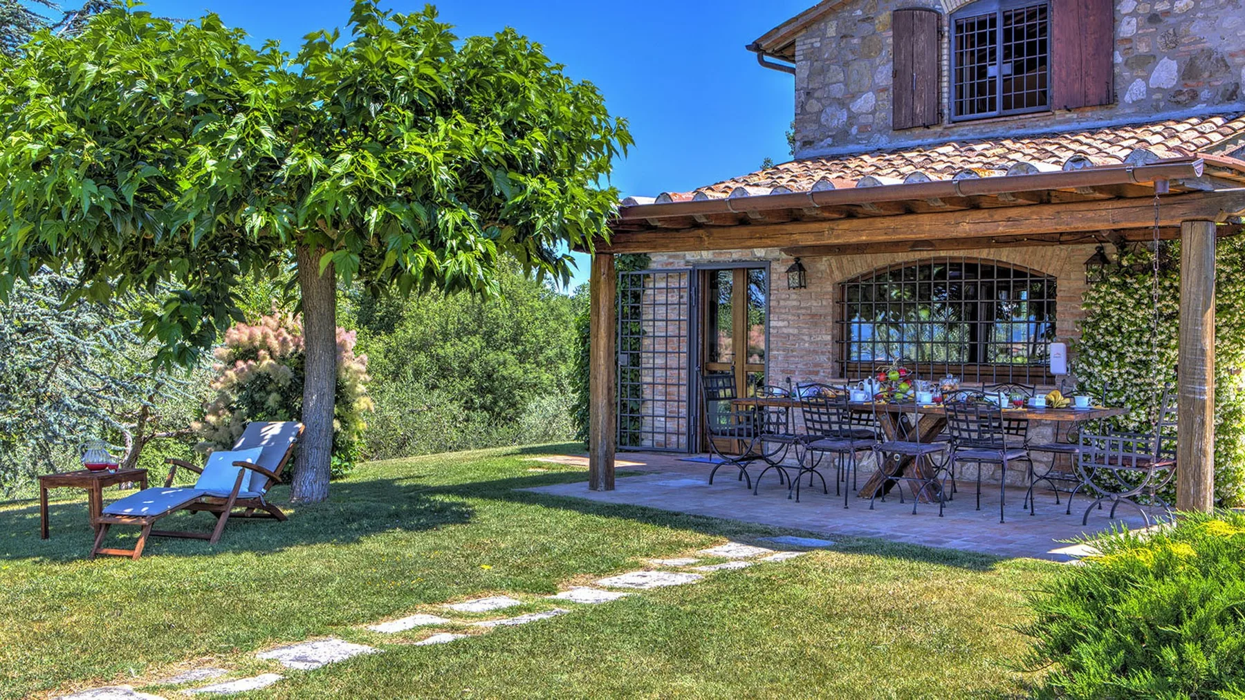 A stone house opens onto a shaded patio with outdoor dining furniture, beside a leafy tree and lawn.