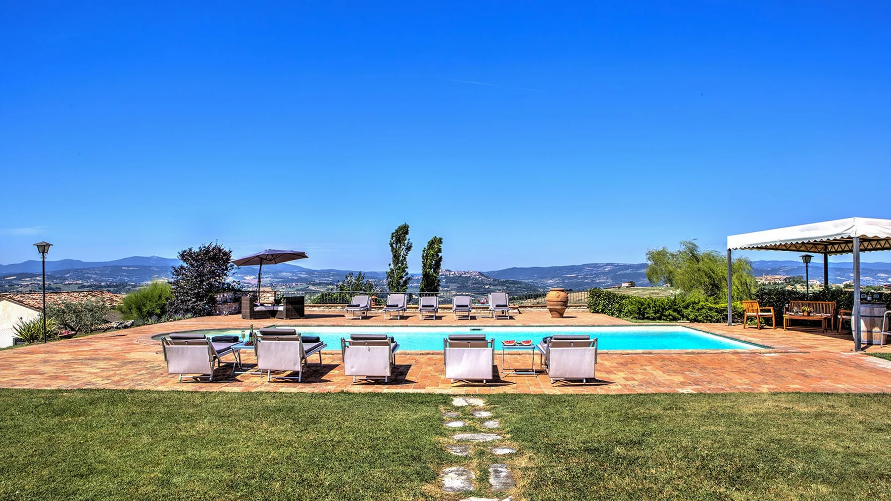 A swimming pool sits in a sunny landscaped yard, with lounge chairs, a pergola, and distant hills.