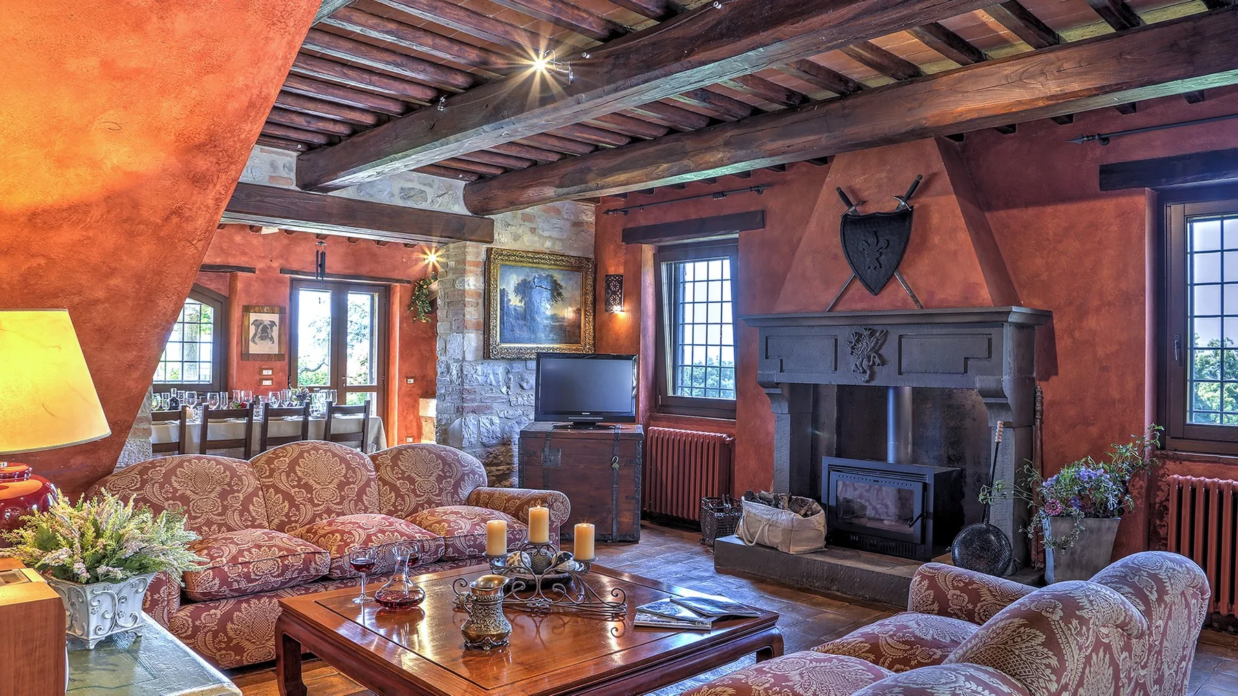 A rustic living room features exposed wooden ceiling beams, patterned sofas, and a large stone fireplace.