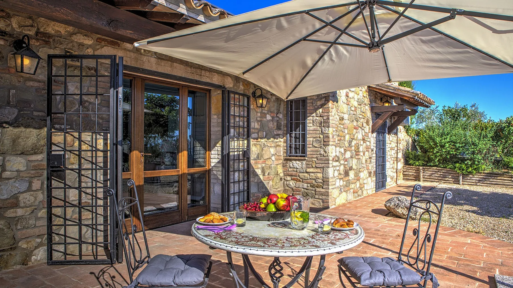 A stone patio has a round table set with fruit and drinks beneath a large umbrella.