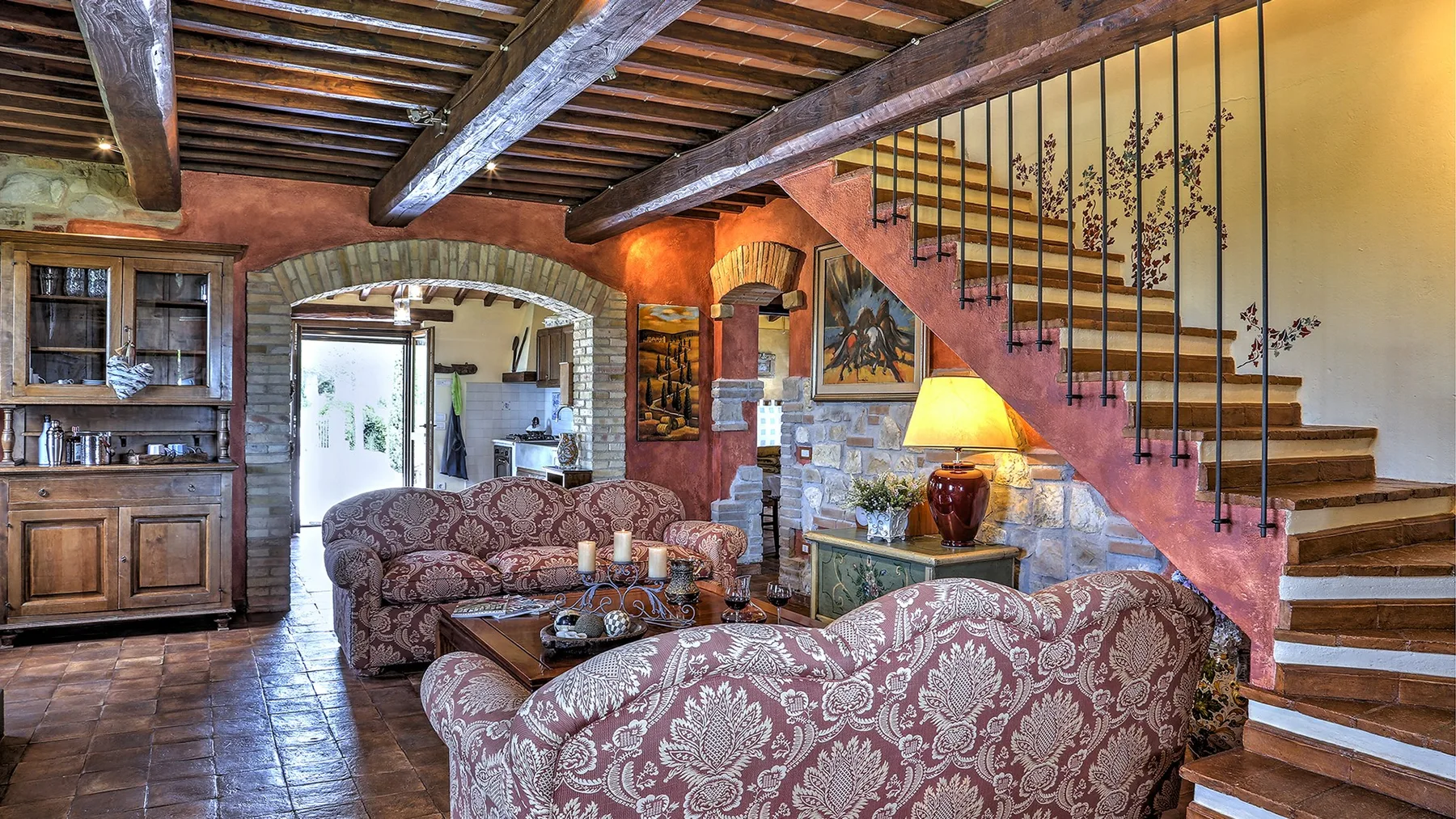 A rustic living room has patterned sofas, a stone fireplace, exposed wooden beams, and a staircase.