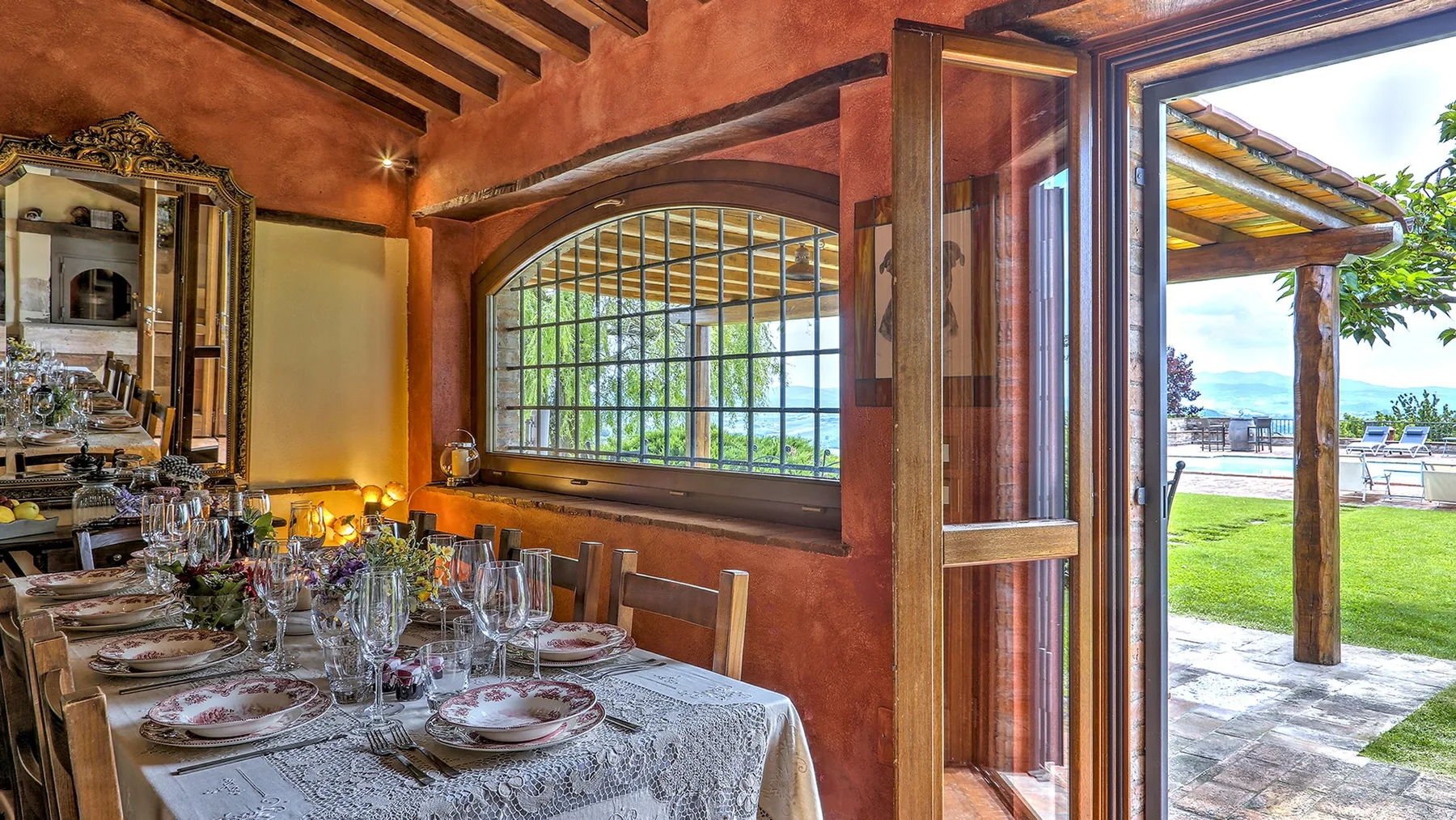 A dining table is set with plates and glassware beside a large arched window and open doors.