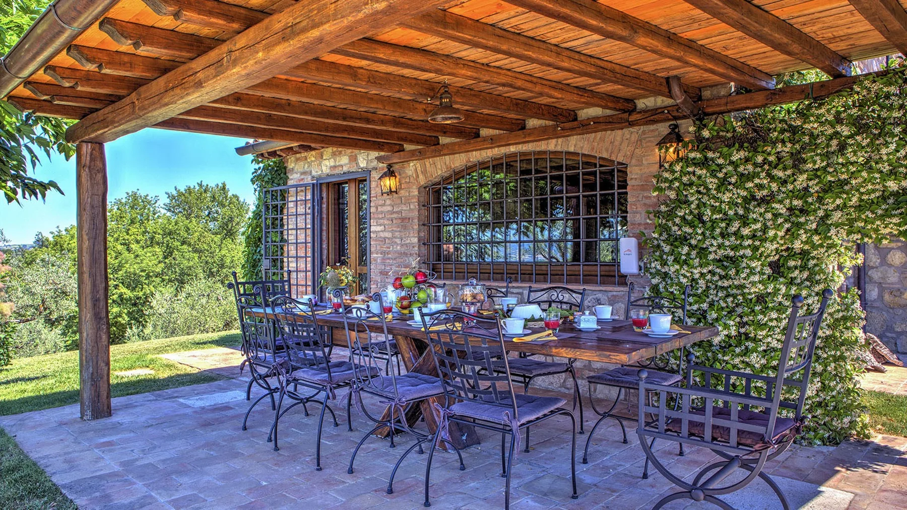A covered stone patio has a long outdoor dining table, metal chairs, and ivy-covered walls.