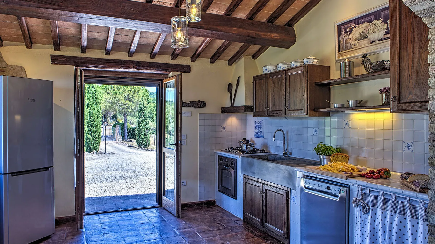 A rustic kitchen has dark wooden cabinets, a tiled backsplash, and open doors overlooking a garden path.