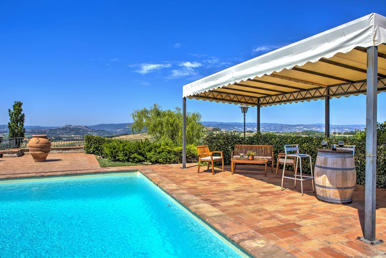 A turquoise swimming pool borders a terracotta patio with a shaded pergola, outdoor seating, and a barrel.