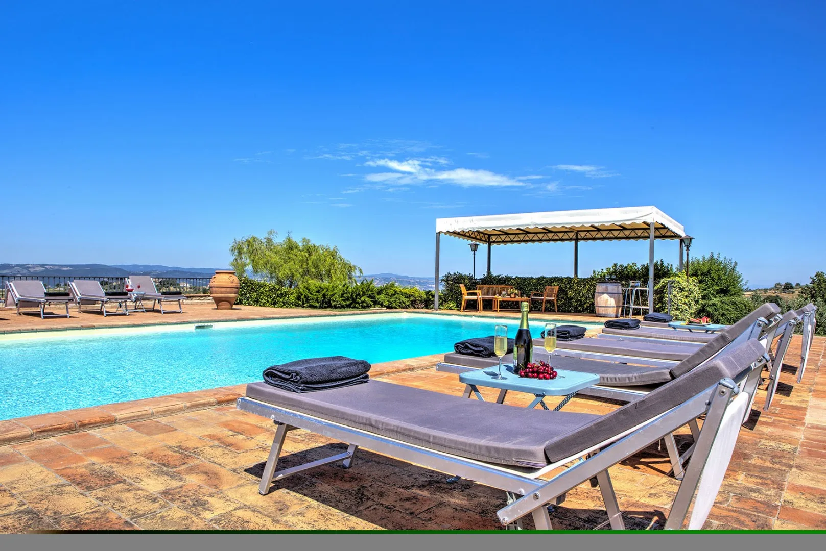 A blue swimming pool is surrounded by cushioned loungers, with a shaded pavilion beyond.